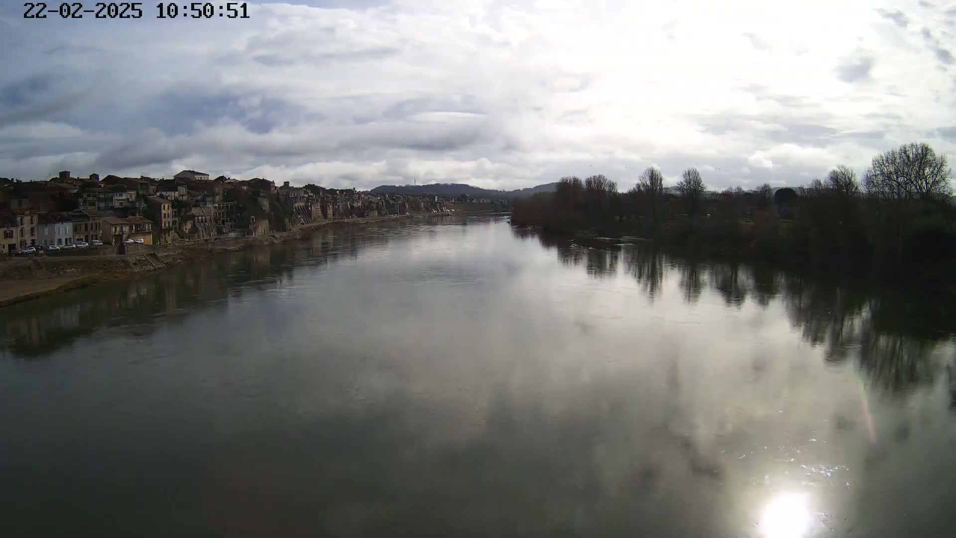 A calm river reflects a cloudy sky and the buildings of a town on its banks, with bare winter trees lining the opposite shore.