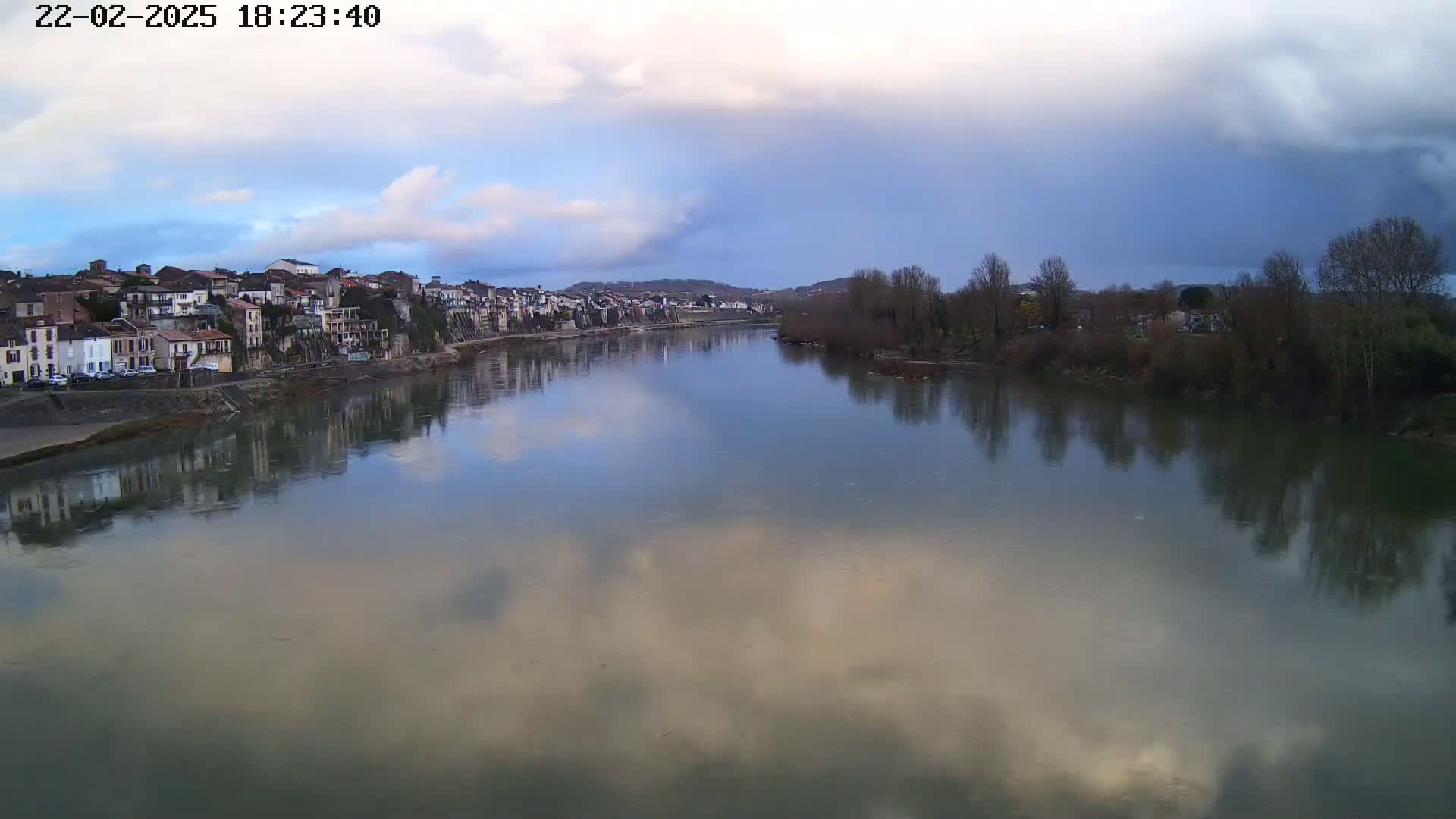 A river reflects a cloudy sky and the buildings of a town on its banks.