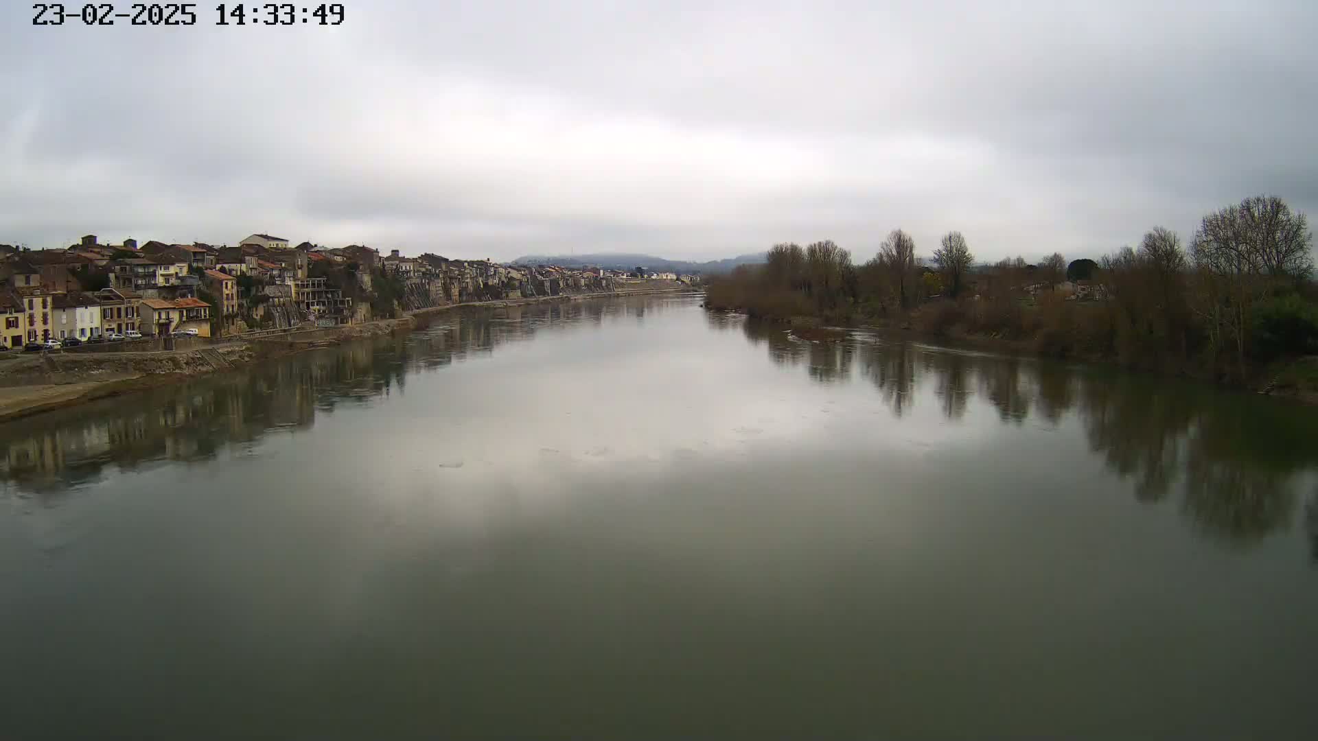 A wide, calm river reflects a row of buildings and bare winter trees under an overcast sky.