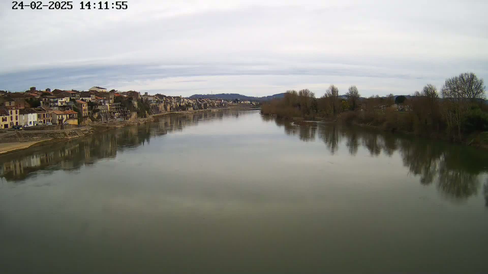 A wide, calm river reflects a town and bare trees under a cloudy sky.