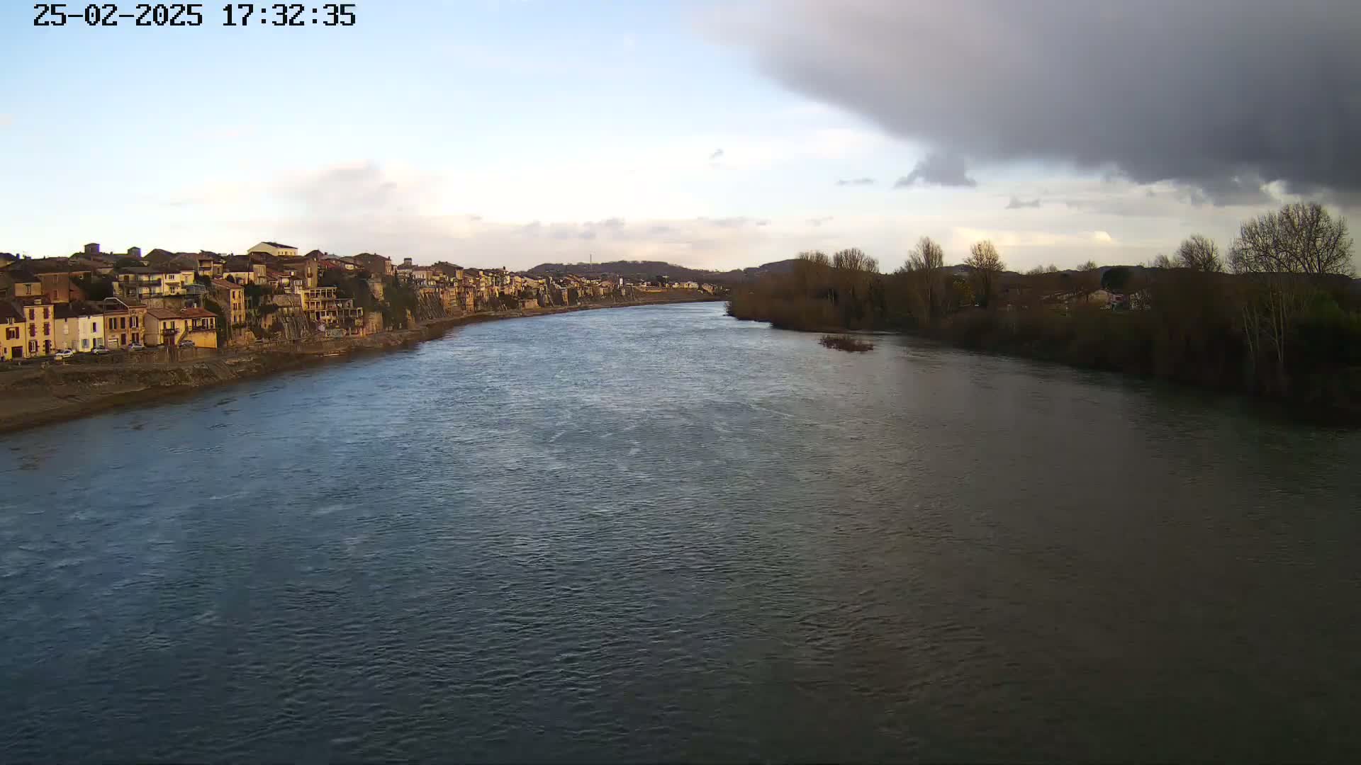 A wide river flows past a town of buildings along one bank and bare trees along the other, under a partly cloudy sky.