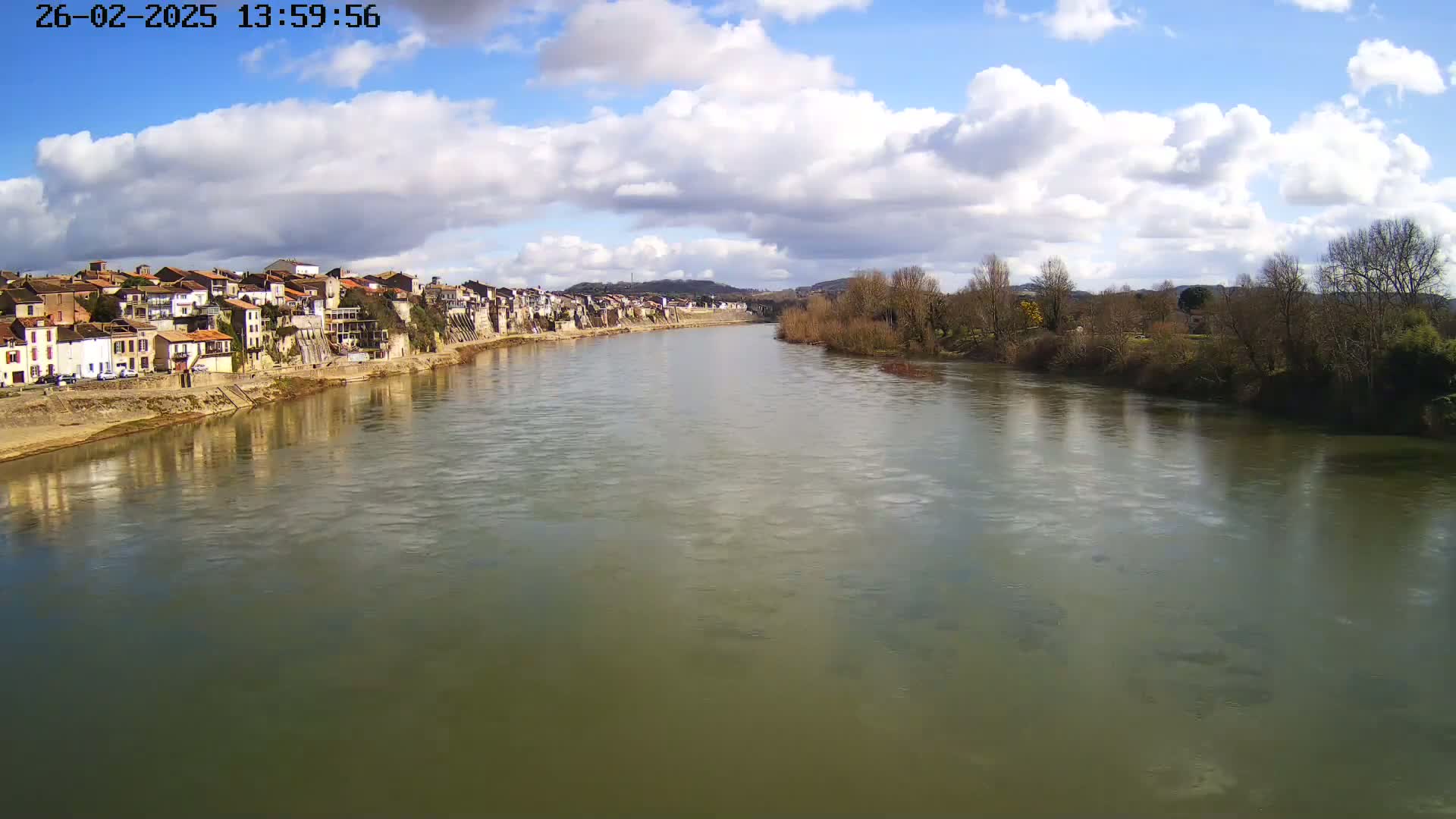 A wide river flows under a partly cloudy sky past a town of closely-packed buildings situated along its bank.