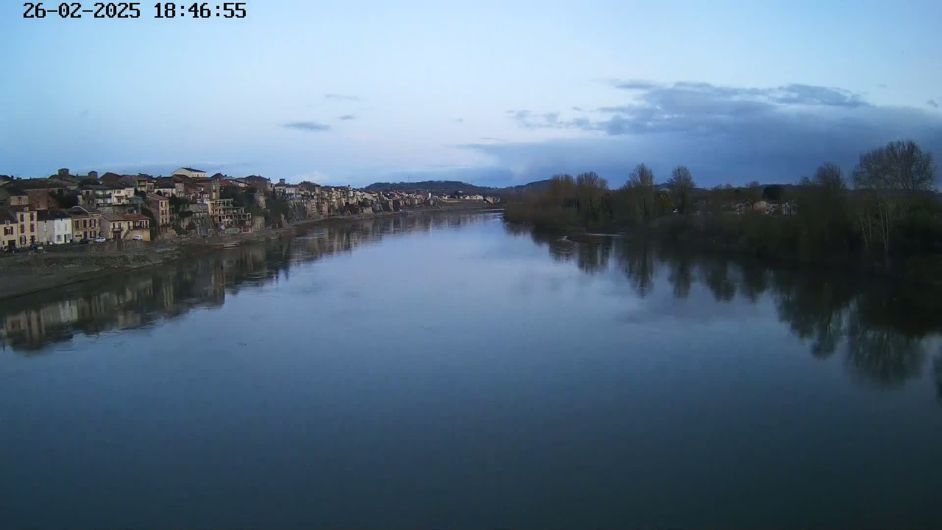 A calm river reflects a town and bare winter trees under a twilight sky.