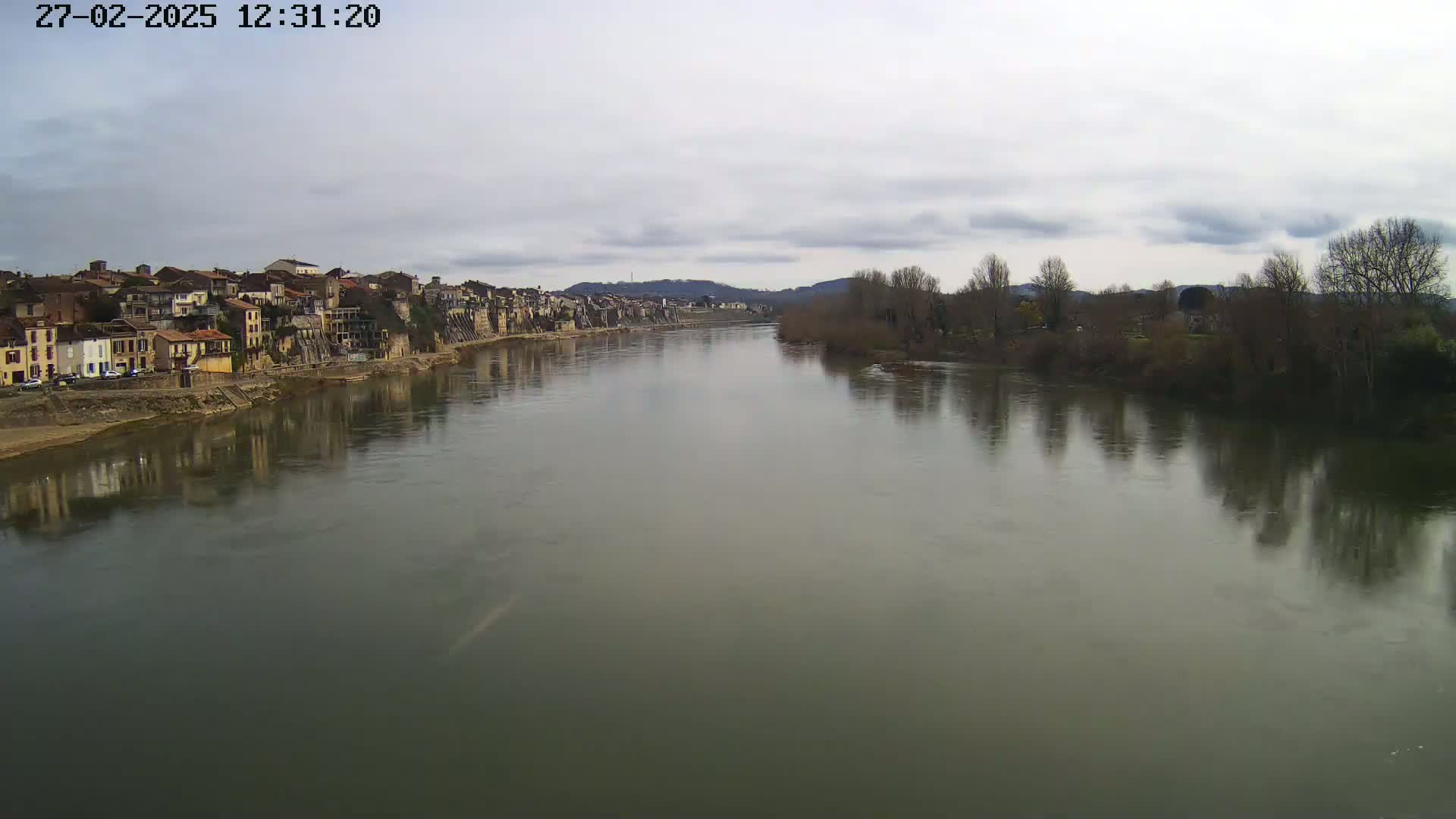 A wide, calm river reflects a town and bare trees under a cloudy sky.