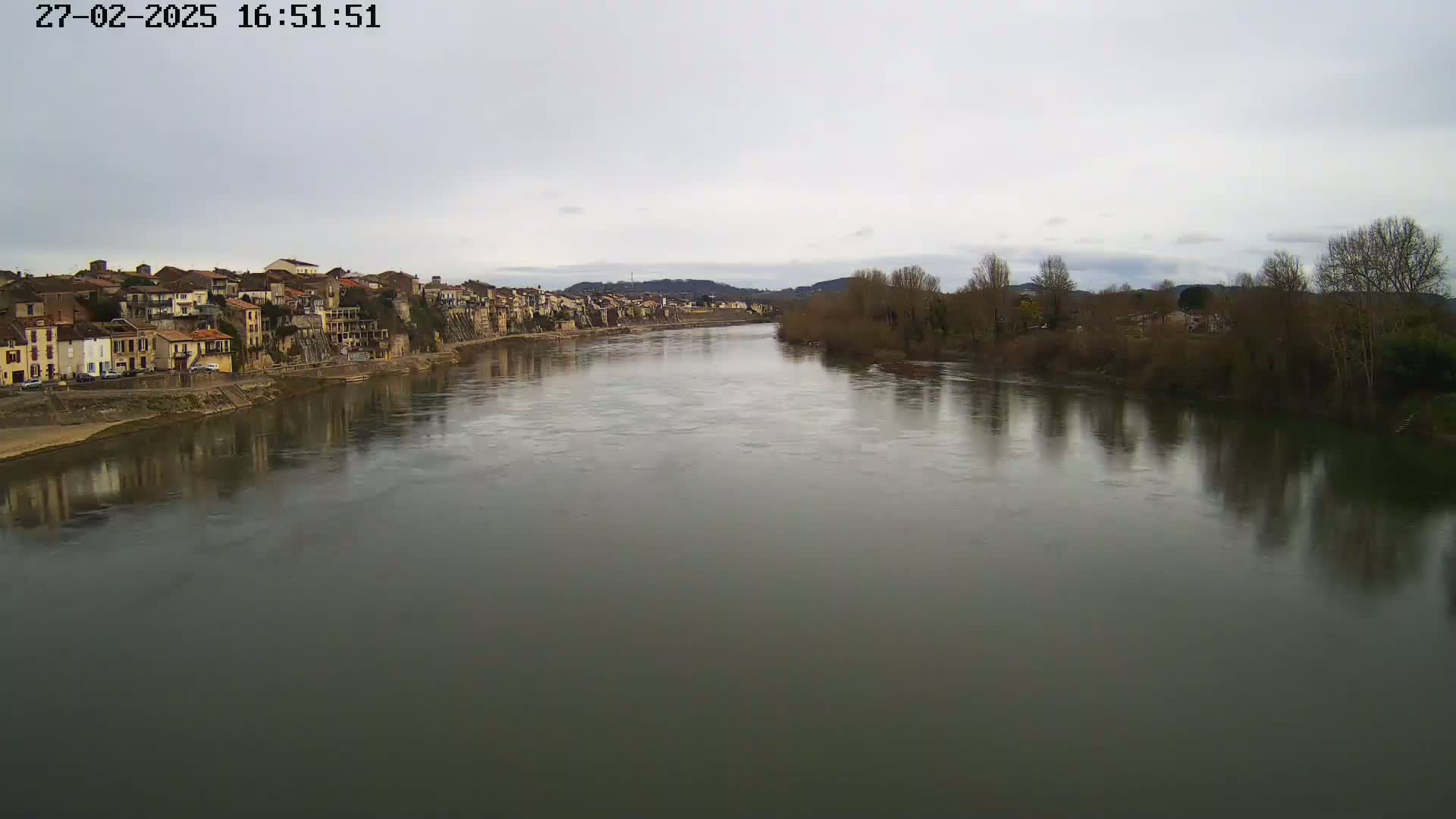 A wide river reflects a town of muted-color buildings and bare winter trees under a cloudy sky.