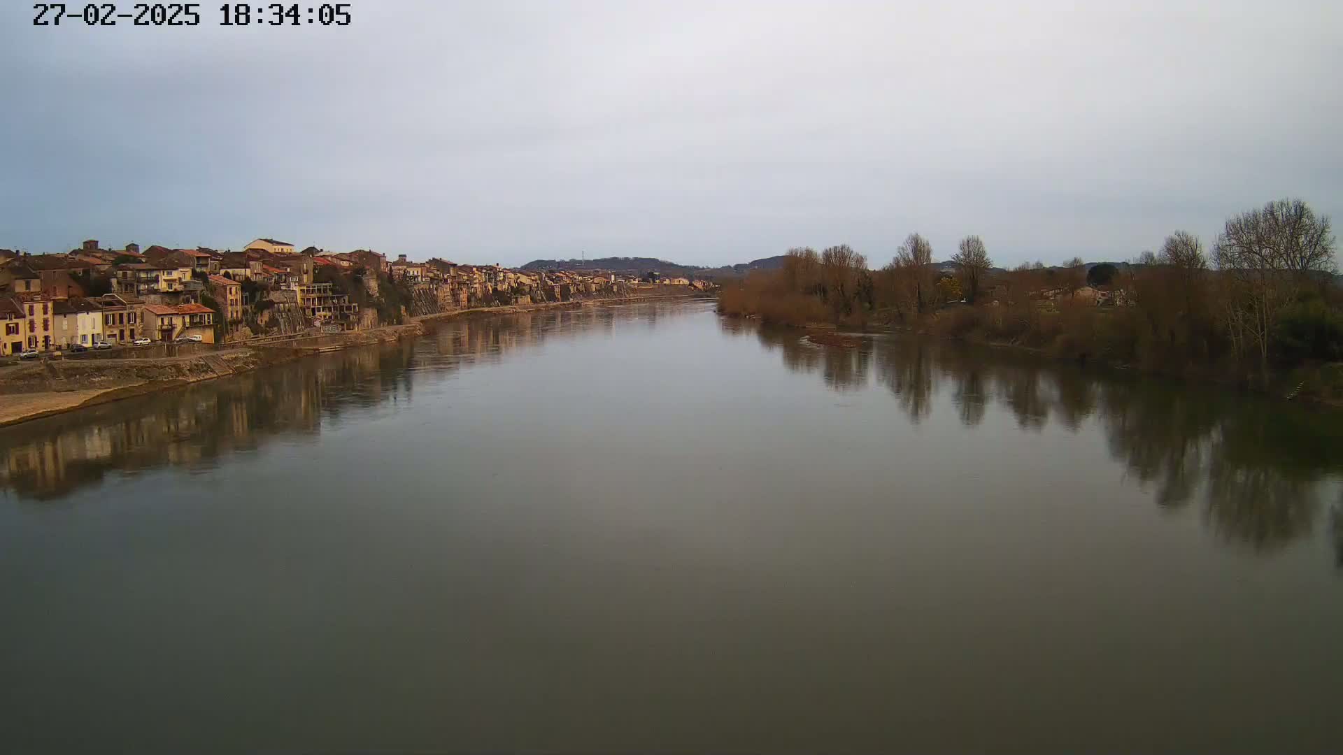A wide, calm river reflects a town of muted, earth-toned buildings and bare winter trees under a pale, overcast sky.