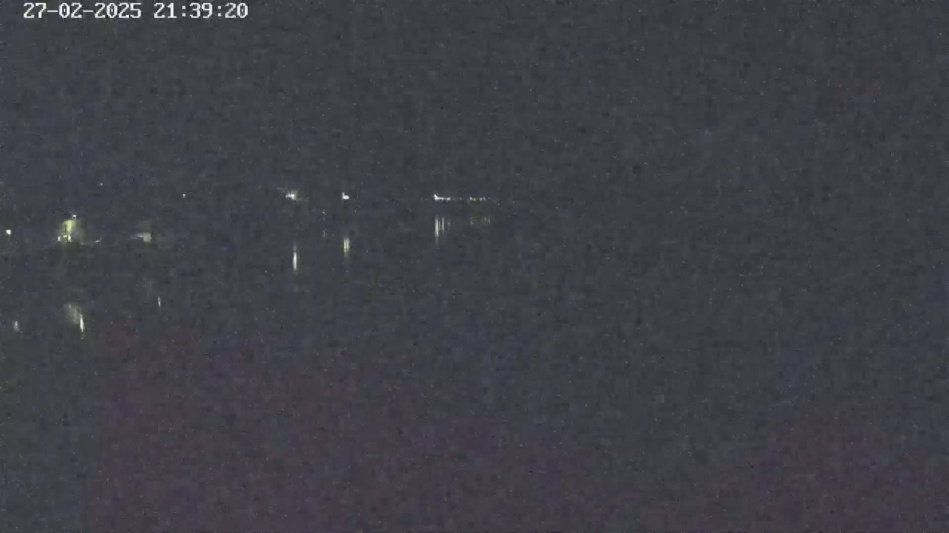 A nighttime view of a dark body of water reflecting faint lights from a distant shoreline under a clear, dark sky.
