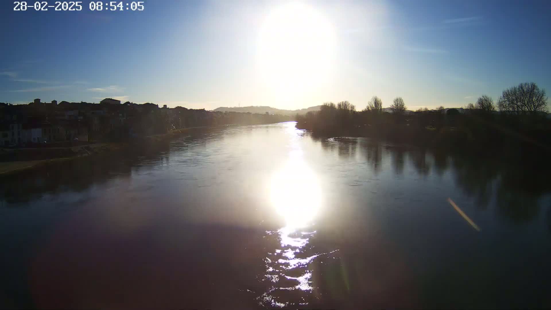 A sunlit river flows past a town on a clear day, reflecting the bare trees and buildings lining its banks.