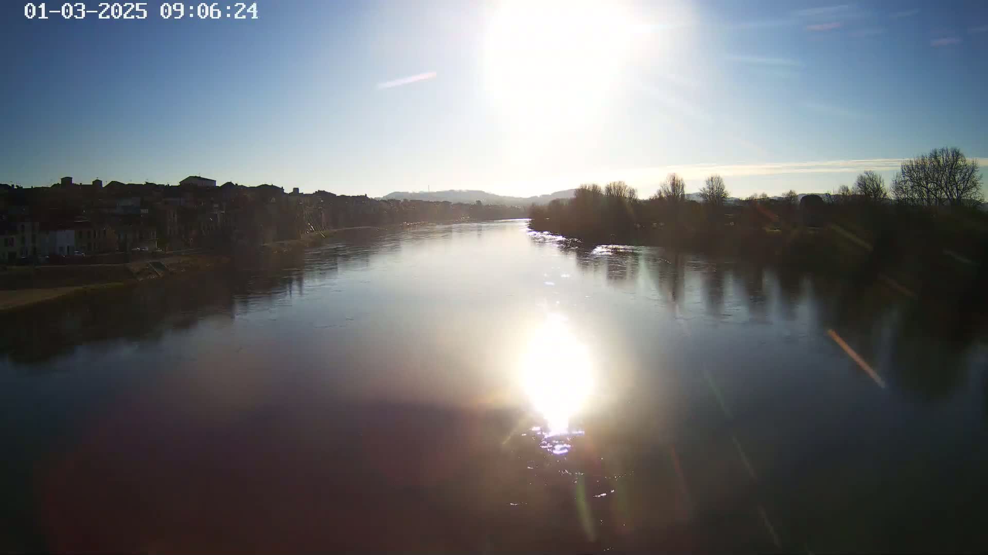 A bright sun shines over a calm river flowing past a town on a clear day.