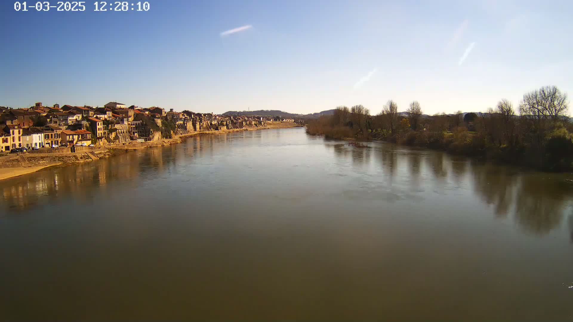 A calm river reflects a town's buildings and bare winter trees under a clear blue sky.