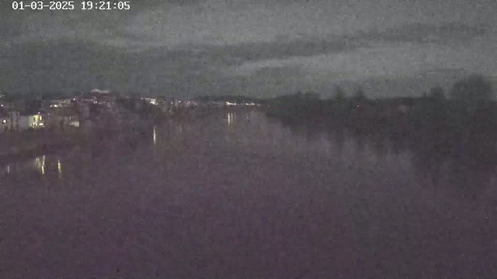 A wide shot at night shows a dark river reflecting the faint lights of a town on the opposite bank under a cloudy sky.