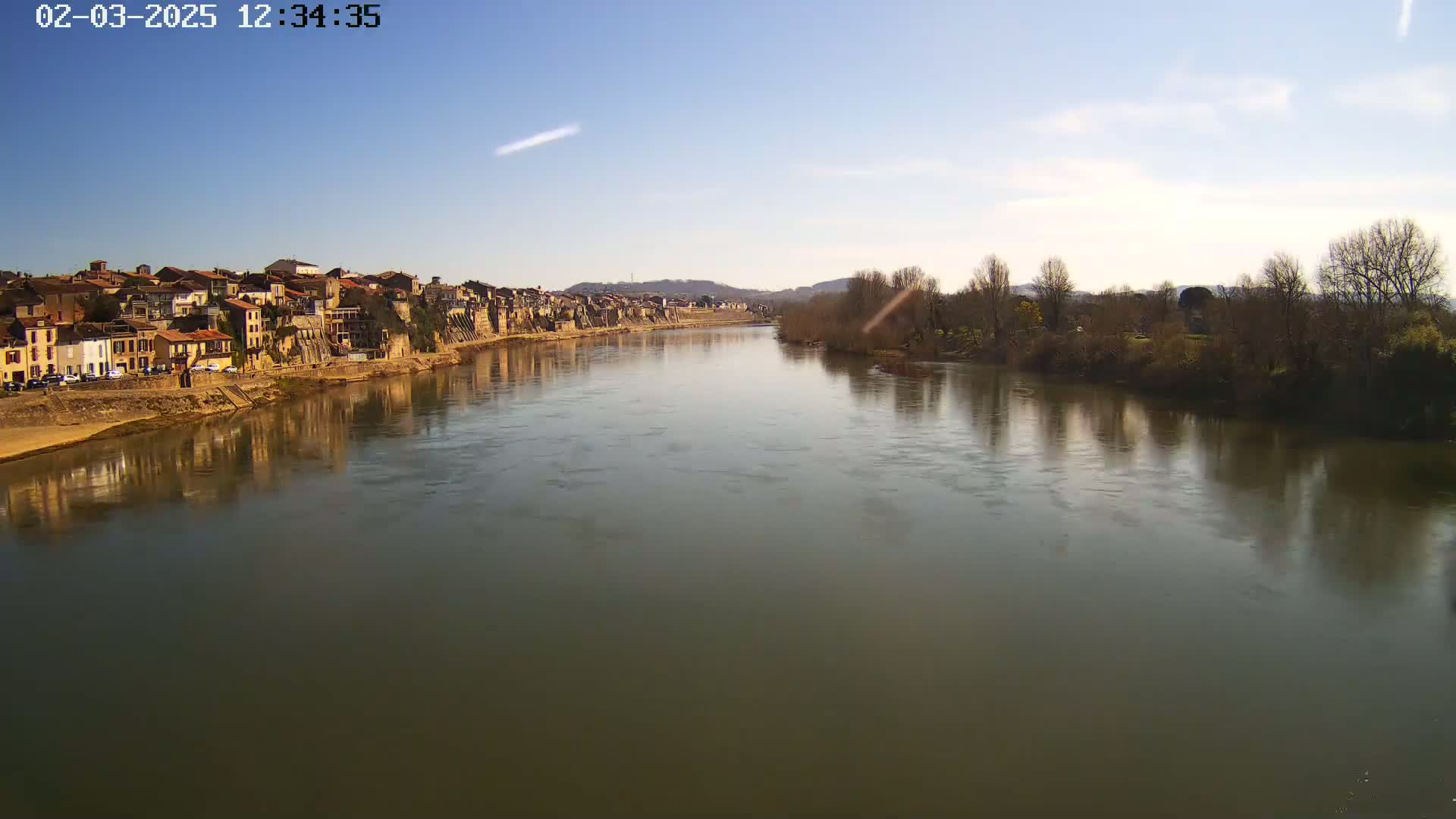 A river reflects a town's buildings and bare winter trees under a mostly clear, sunny sky.