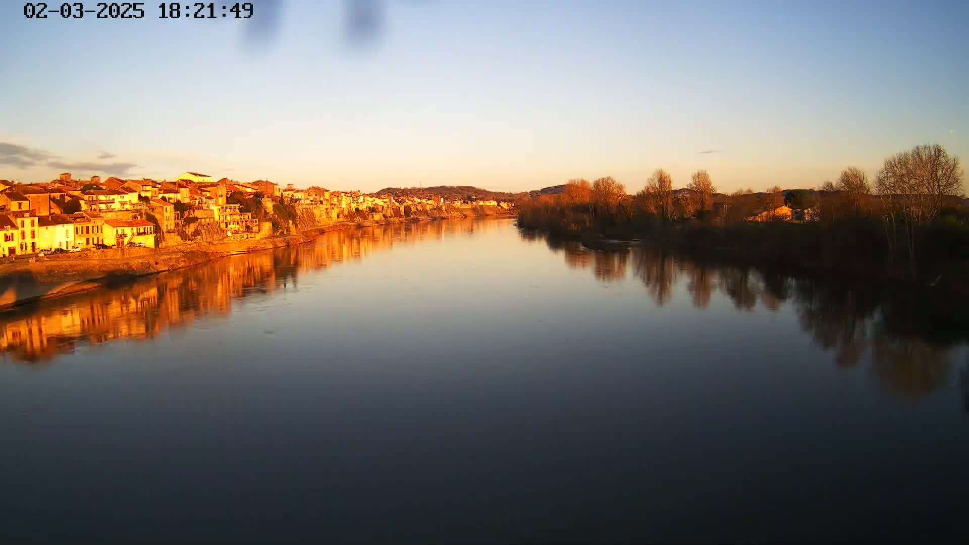 A calm river reflects the setting sun's golden light on a row of buildings and bare winter trees.