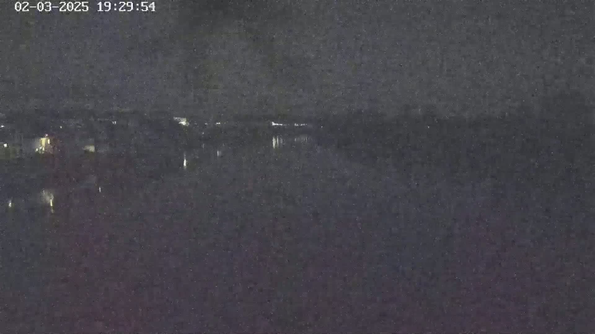 A dark, nighttime view of a body of water reflecting a few distant lights on the opposite shore under a clear sky.