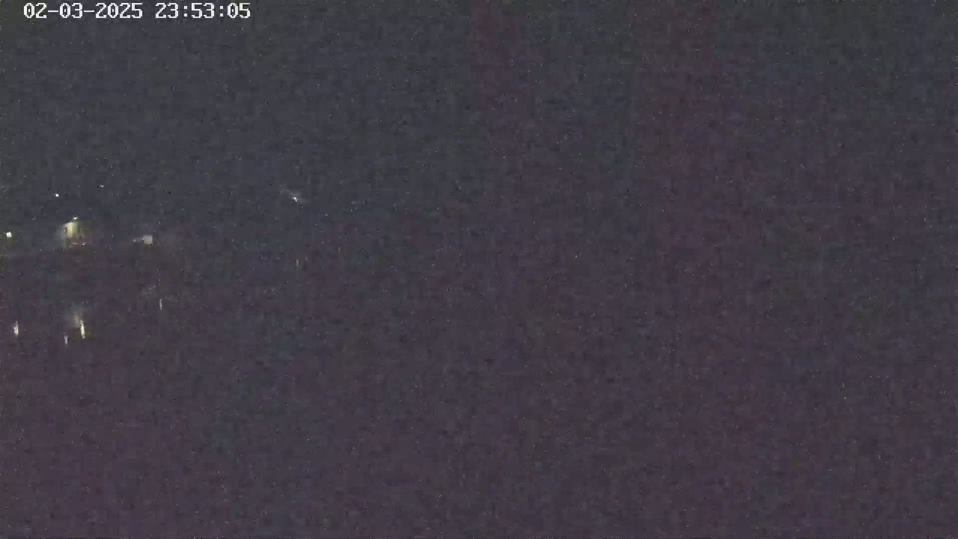 A nighttime, low-light image shows a dark, indistinct landscape with a few faint lights visible in the distance.