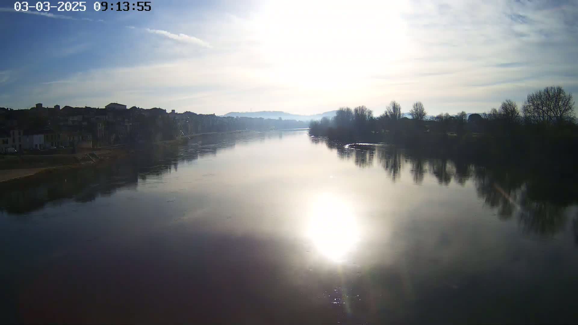 A calm river reflects a bright sun and the silhouettes of buildings and trees on a partly cloudy day.