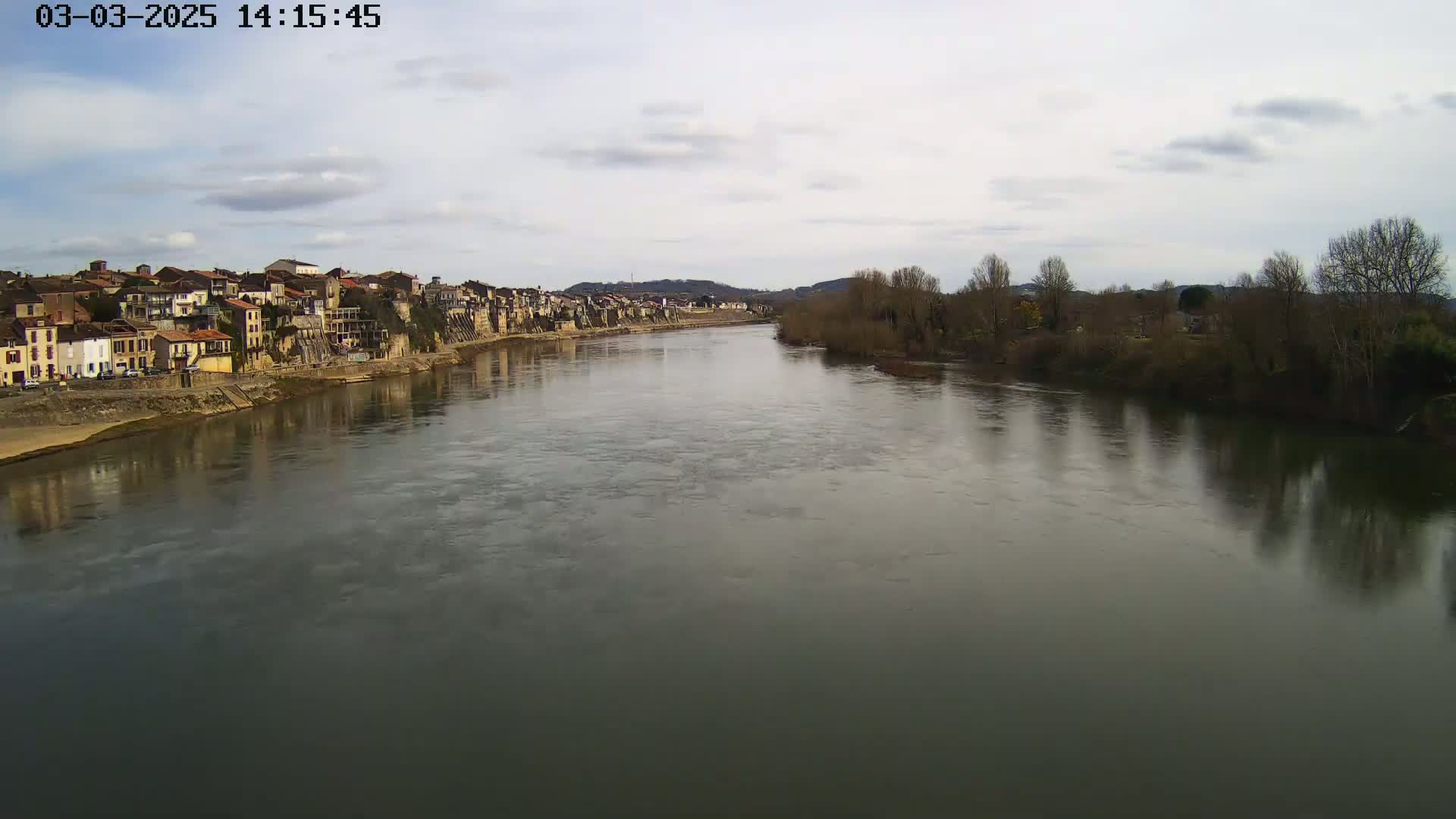 A wide river flows past a town of closely-packed buildings under a mostly cloudy sky.