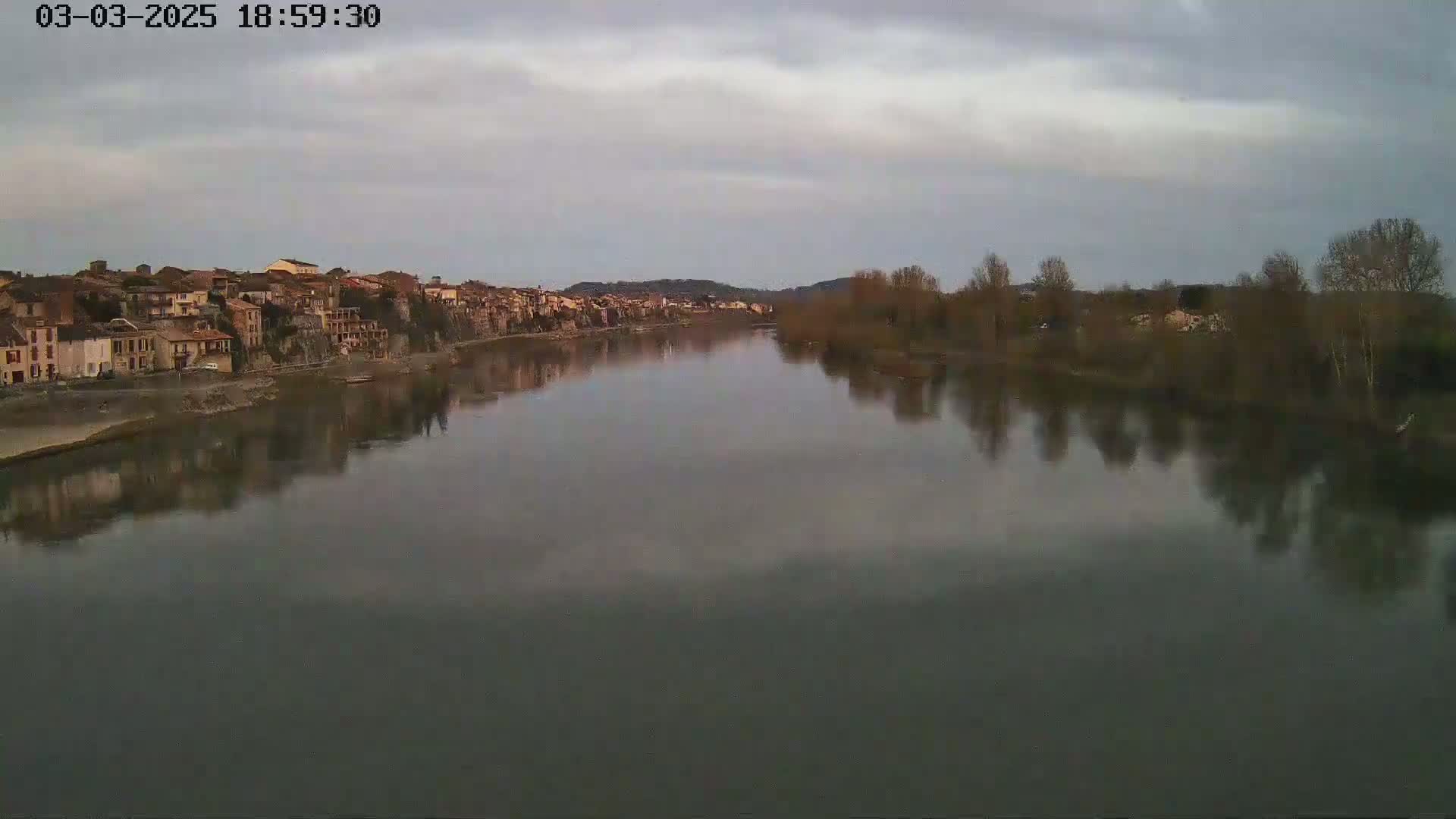A calm river reflects a town of muted brown buildings under a cloudy sky.
