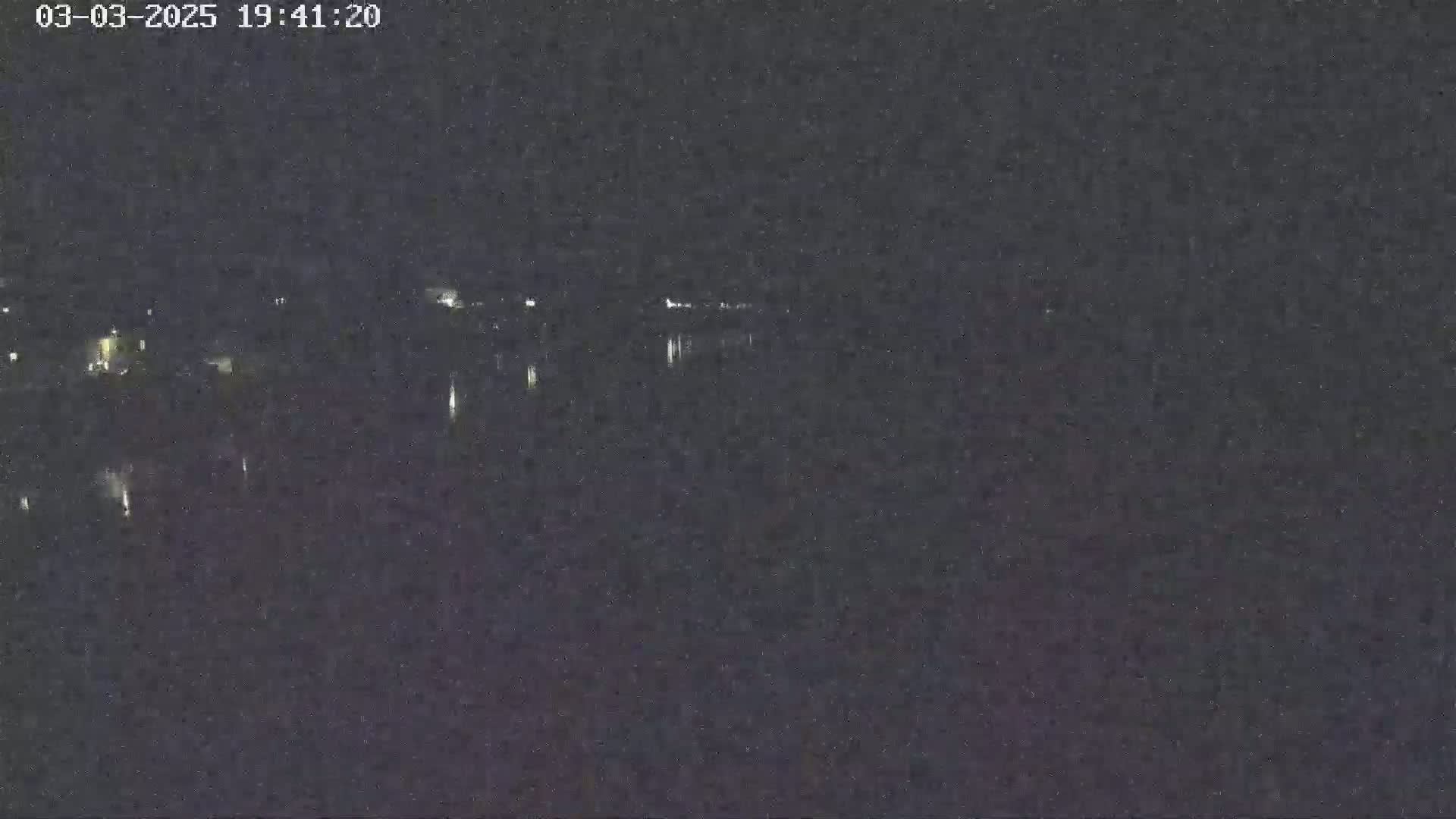 A nighttime long shot shows a dark body of water with faint lights on the far shore under a dark sky.
