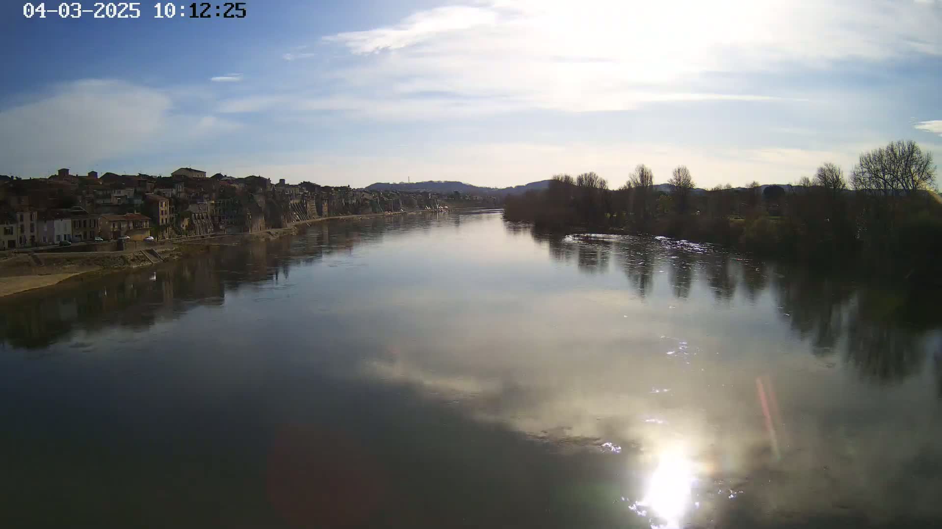 A calm river reflects a partly sunny sky and the buildings and trees lining its banks.