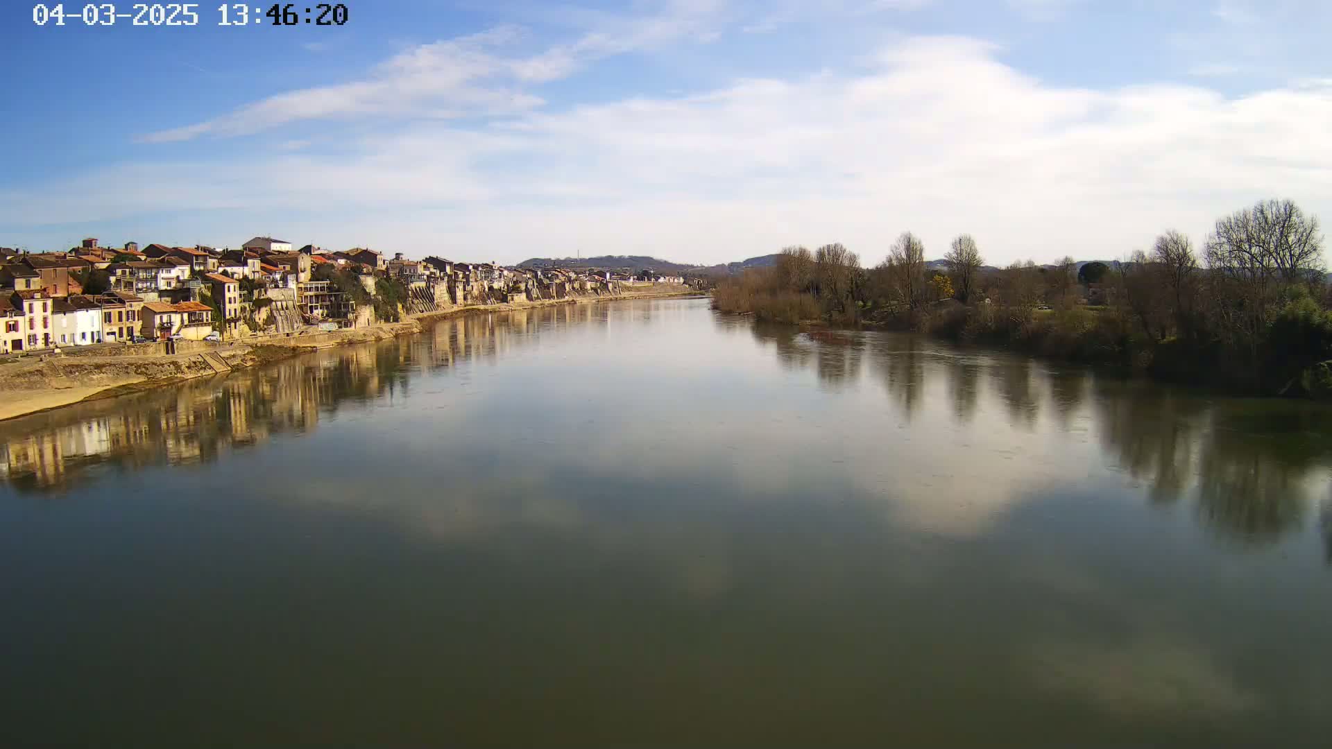 A calm river reflects a partly cloudy blue sky and the buildings of a town on its banks, which are lined with leafless trees.