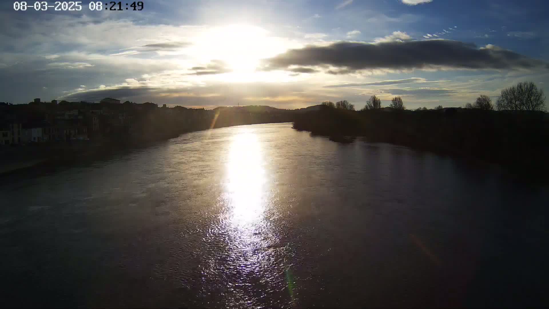 A wide river reflects the setting sun under a partly cloudy sky, with a cityscape visible on one bank and a treeline on the other.