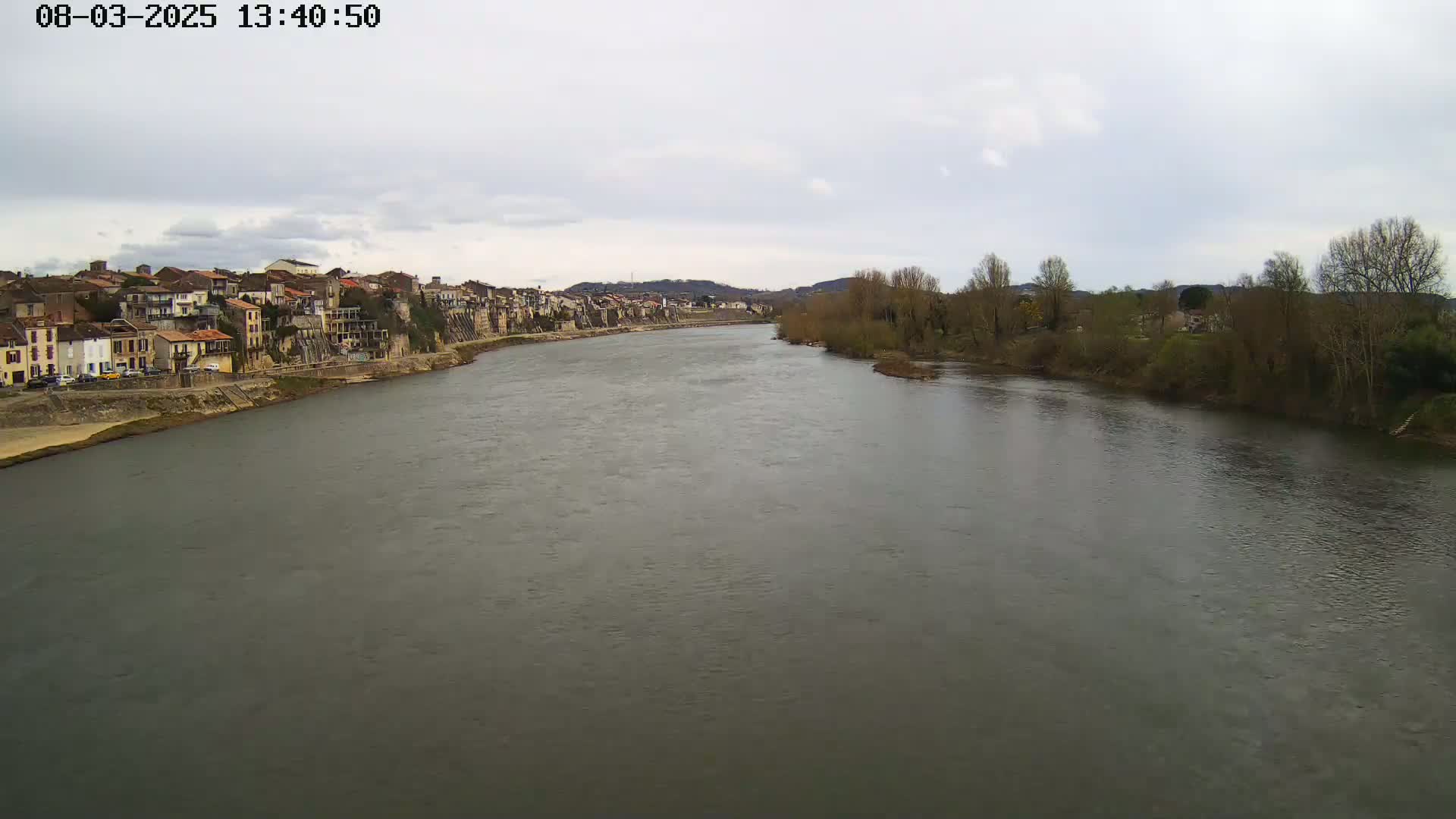 A wide river flows past a town of closely packed buildings under a cloudy sky.