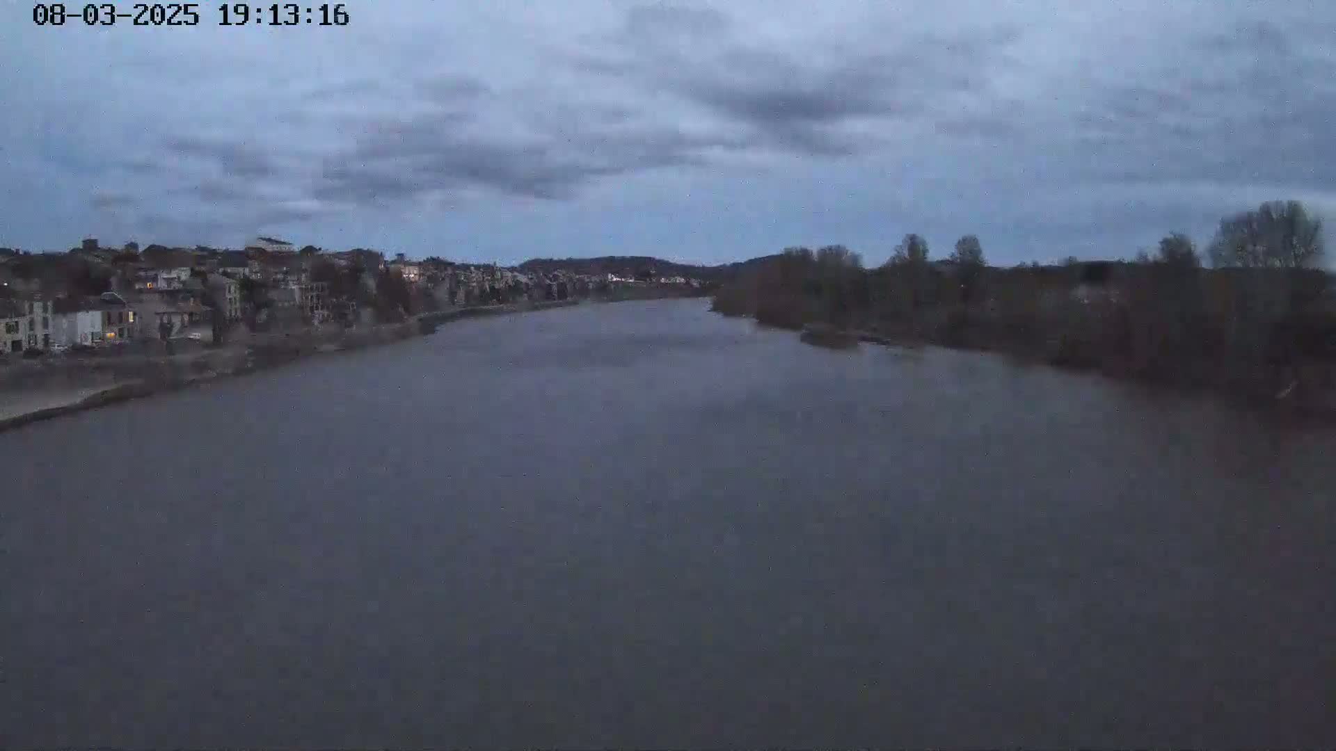 A wide, dark river flows past a town under a cloudy, twilight sky.