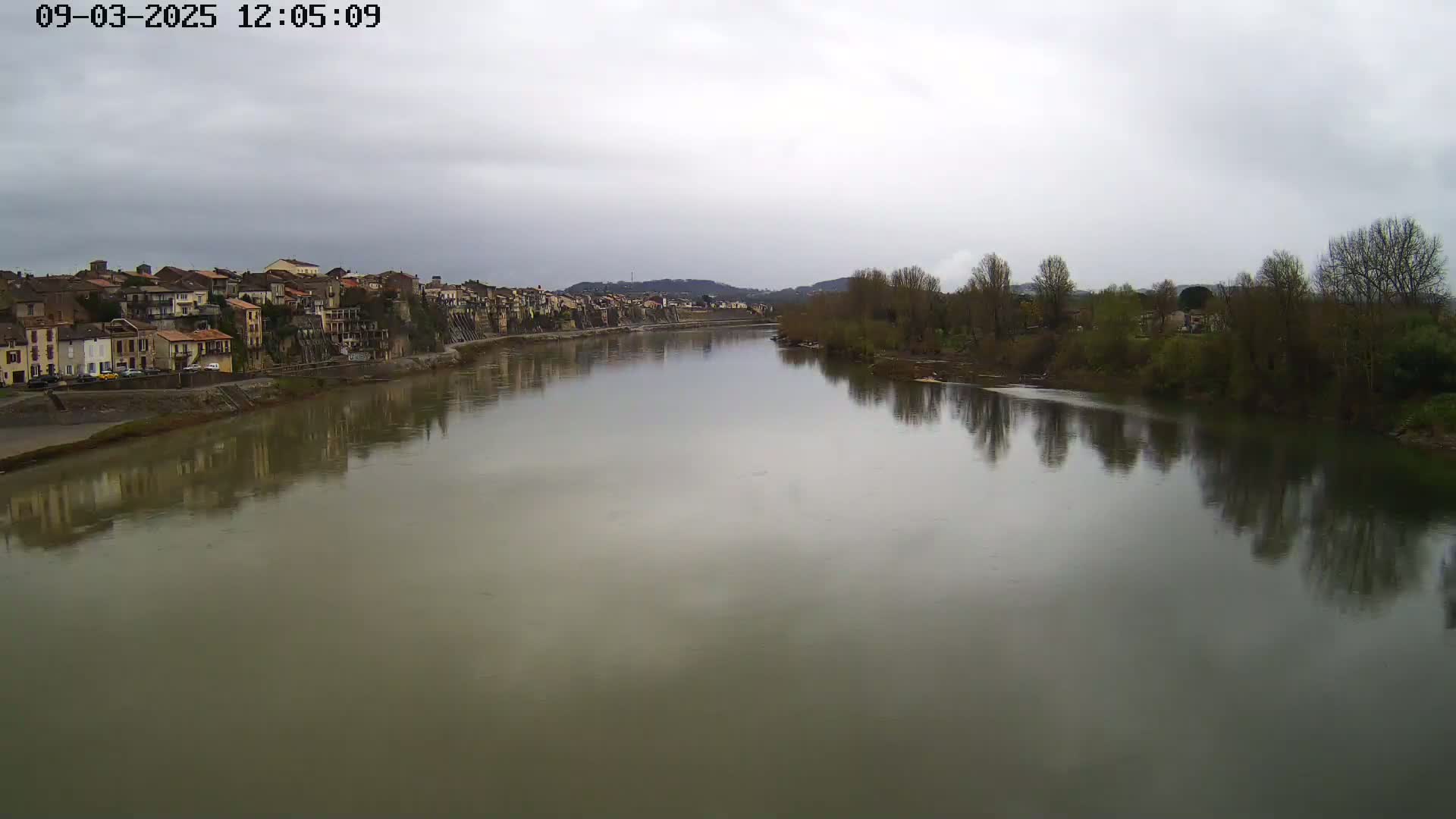 A wide, calm river reflects a town and bare trees under an overcast sky.