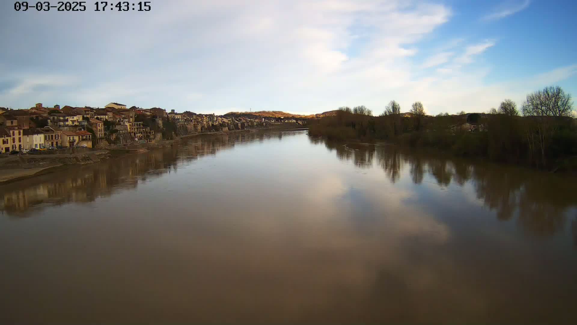 A wide, calm river reflects a partly cloudy sky and the silhouettes of bare winter trees and a town on its banks.