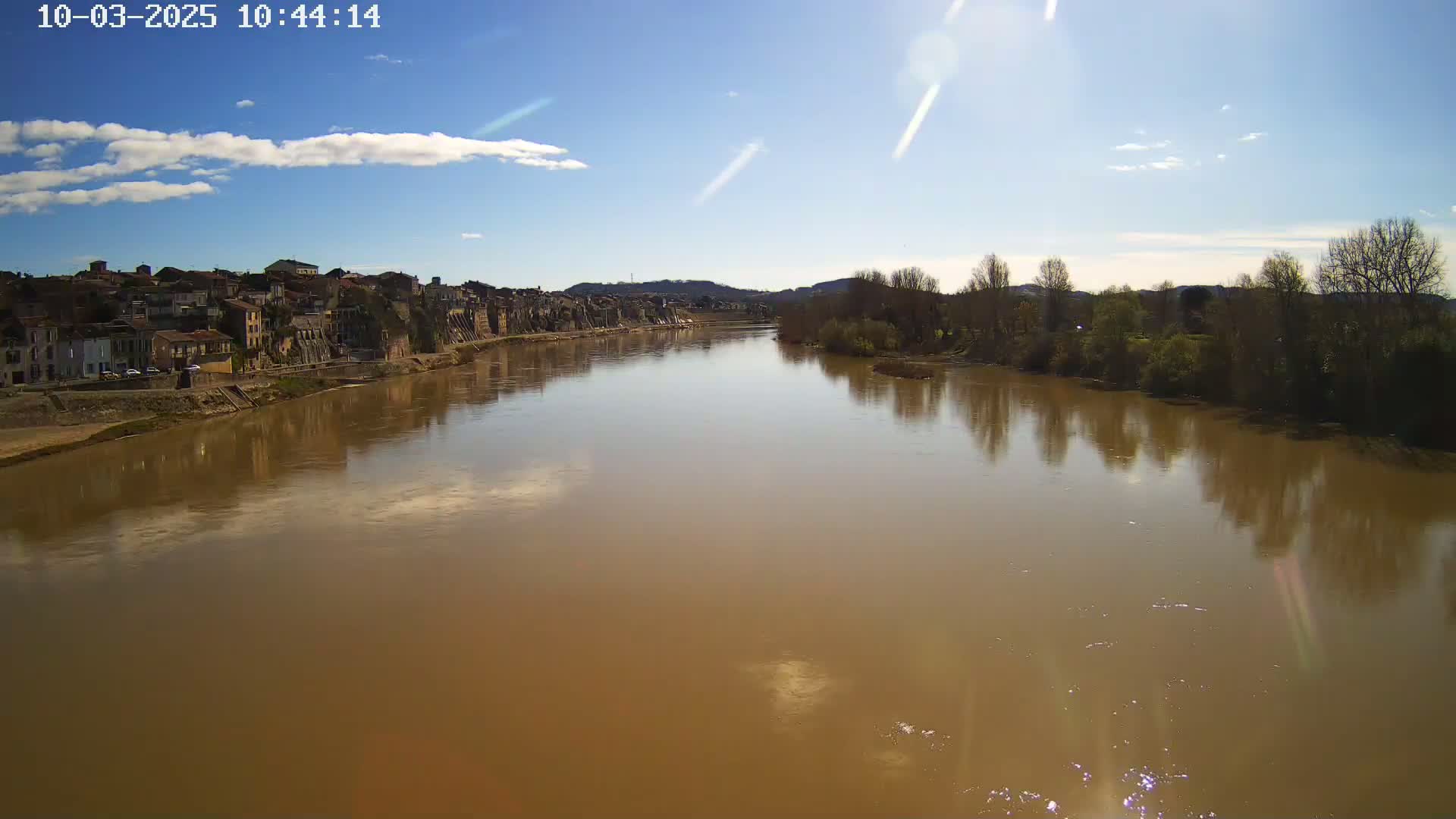 A wide, muddy river reflects a partly sunny sky and the buildings and trees along its banks.