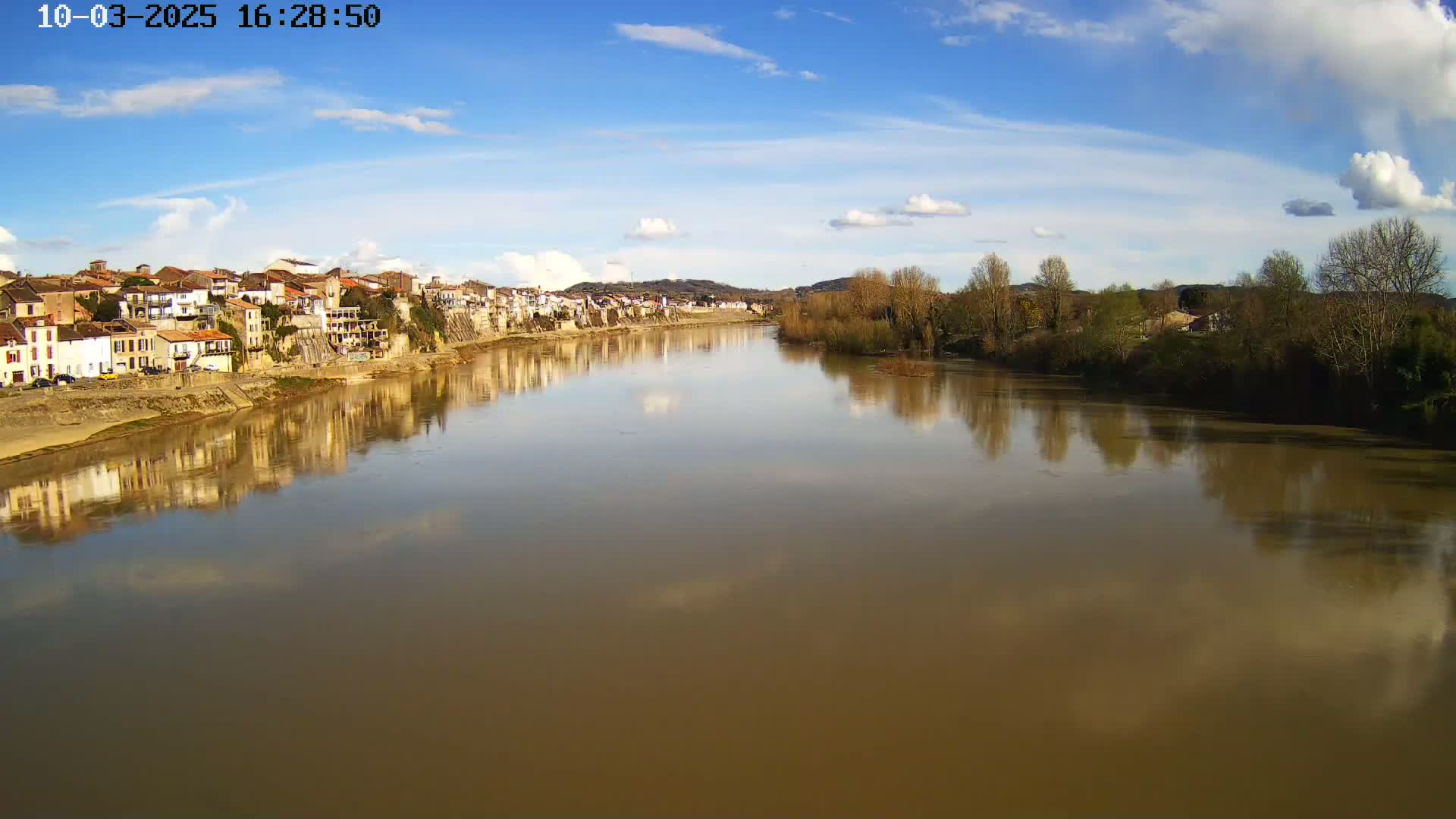A river with muddy-brown water reflects a town's buildings under a partly cloudy, sunny sky.