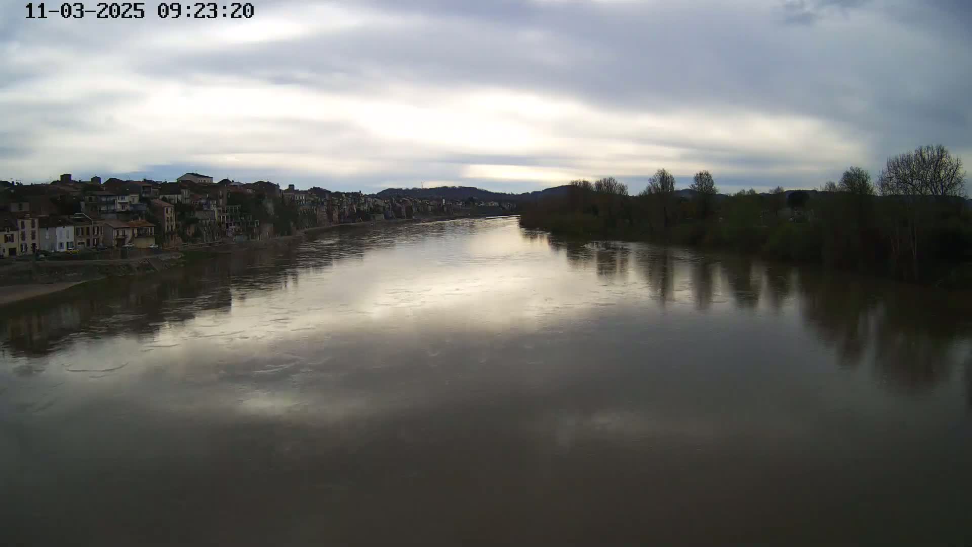 A river reflects a cloudy sky and the silhouettes of buildings and trees on its banks.