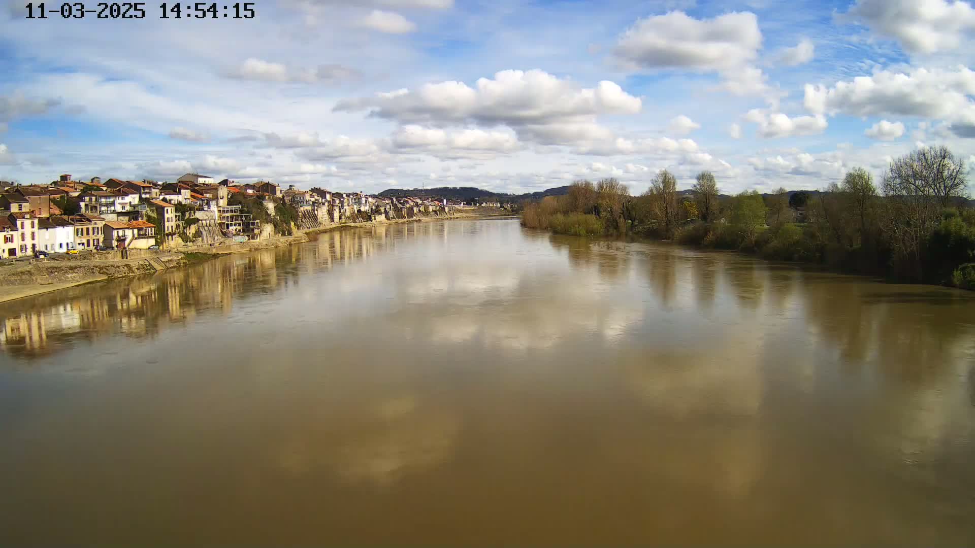 A wide, muddy river reflects a partly cloudy sky and the buildings of a town situated on its bank.