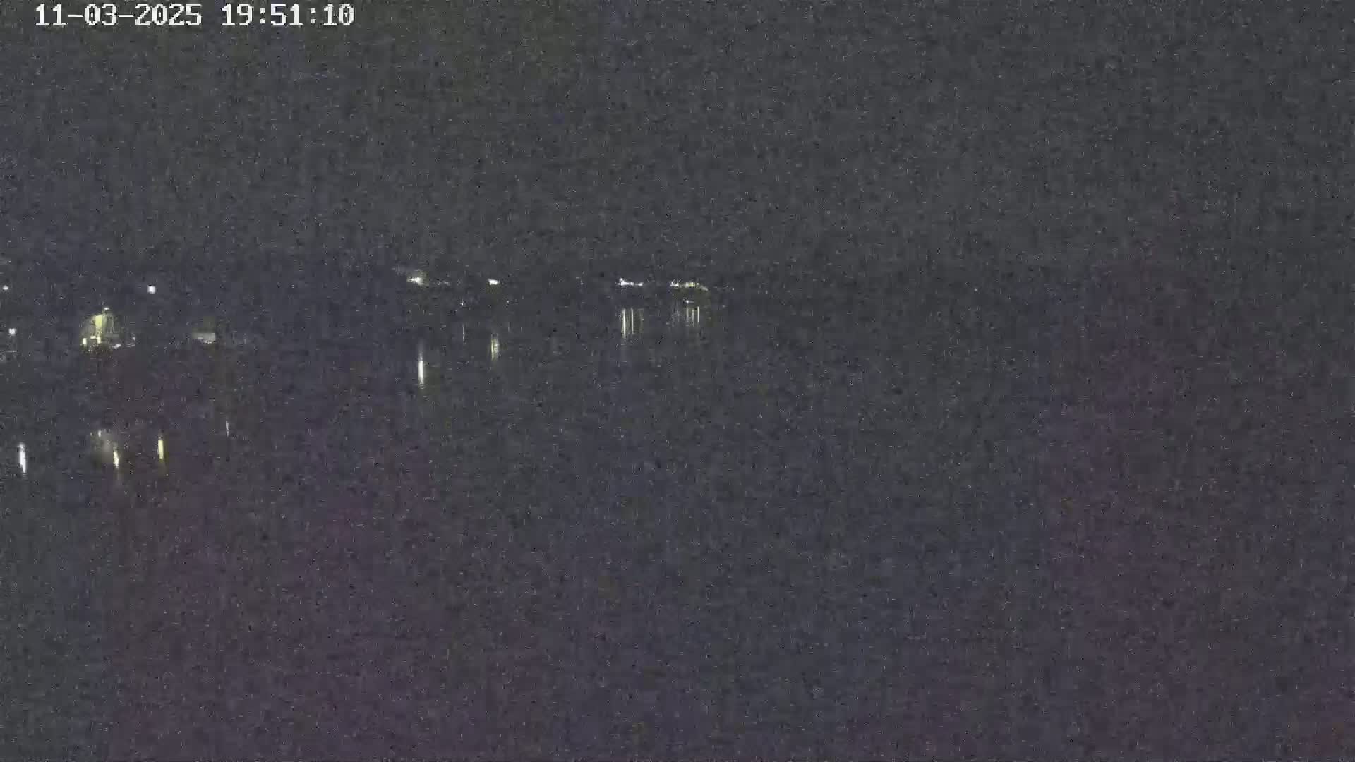 A nighttime view of a calm body of water reflecting dim lights from a distant shoreline under a dark sky.