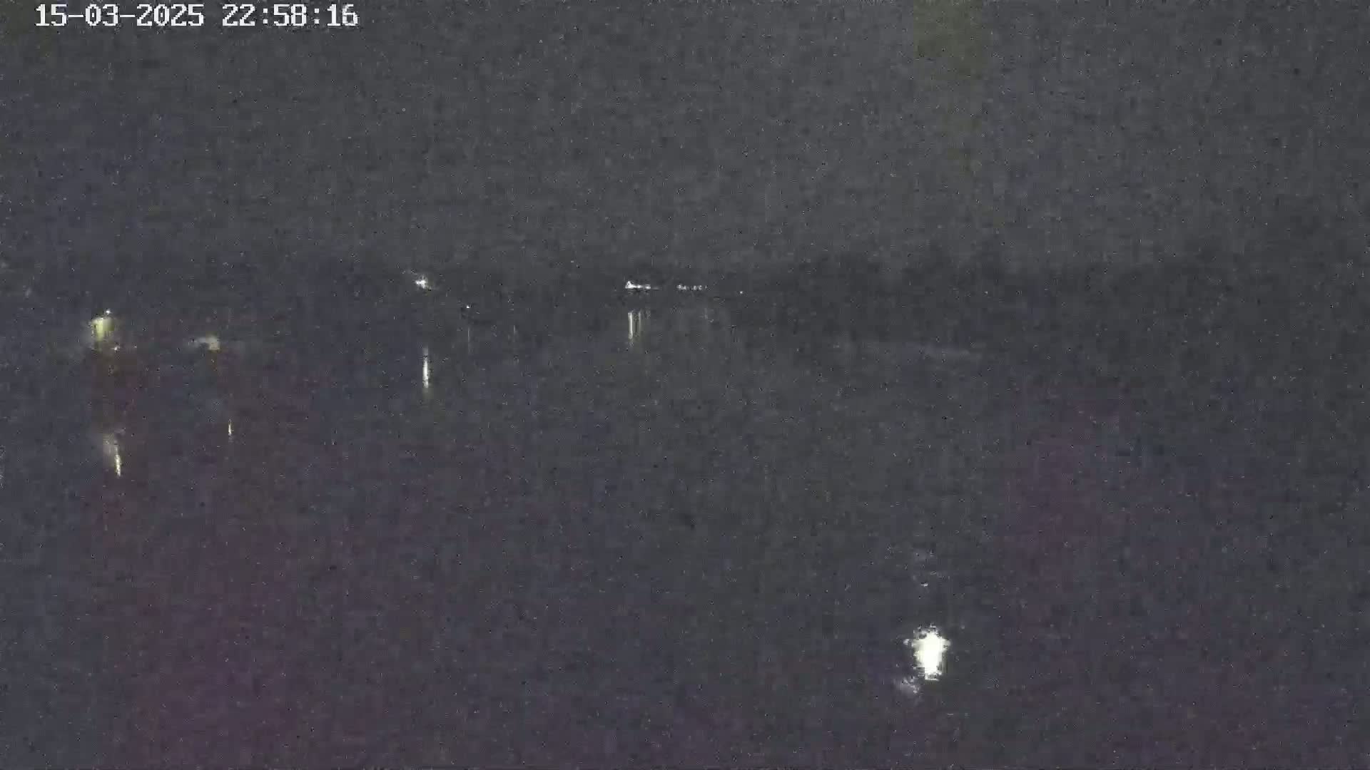 A nighttime, low-light view shows a dark body of water with a few small, distant lights reflecting on its surface.