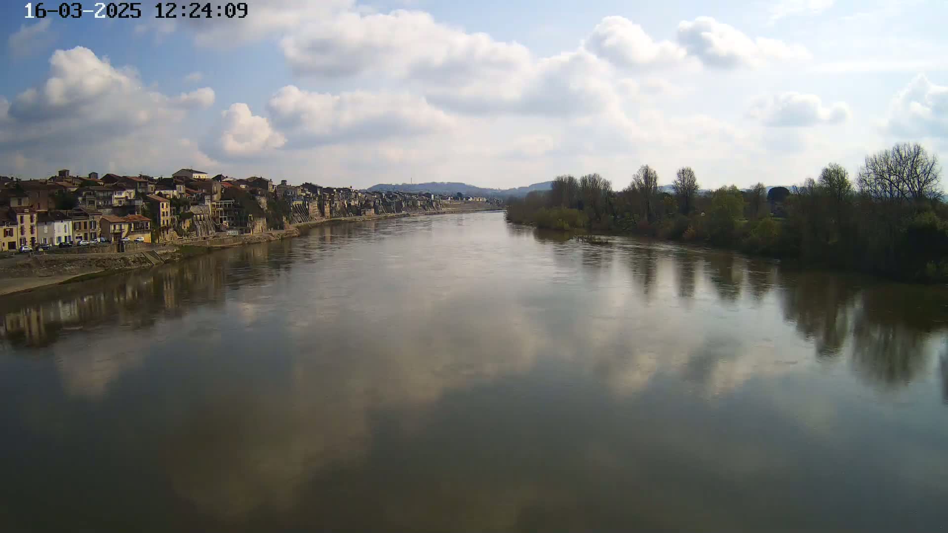 A wide, calm river reflects a partly cloudy sky and the buildings of a town on its bank, with bare trees lining the opposite shore.