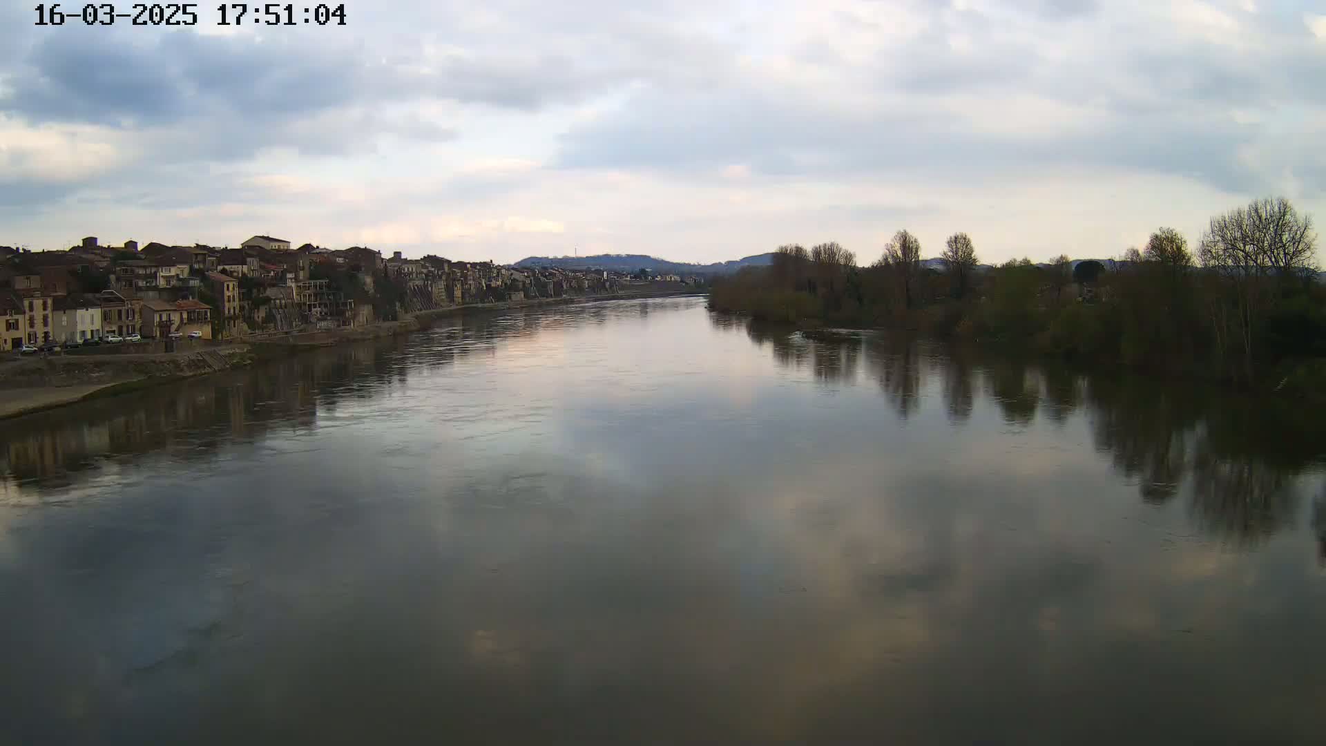 A calm river reflects a cloudy sky and the buildings of a town on its banks, with bare trees lining the opposite shore.