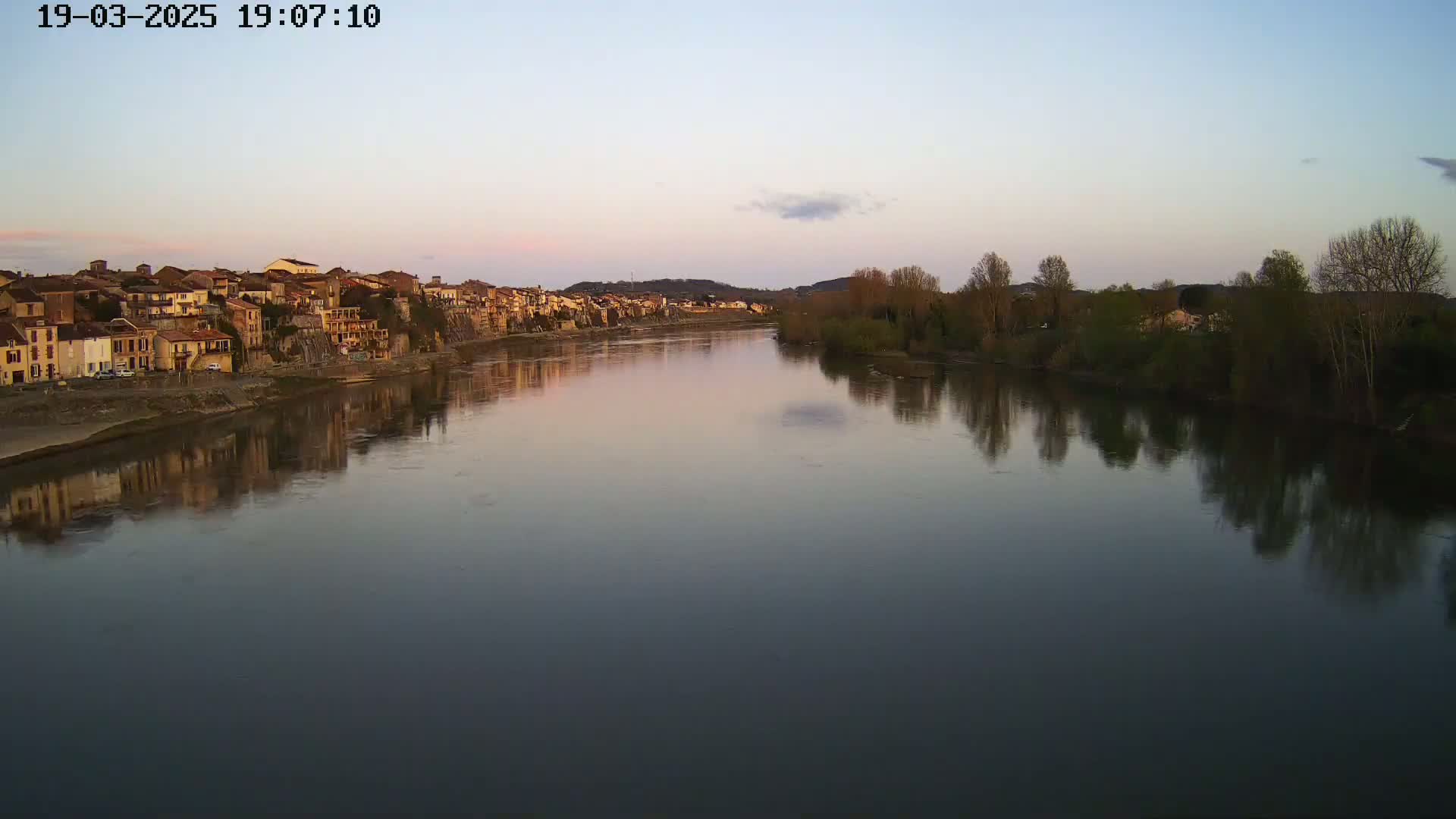 A calm river reflects the pale evening sky and a line of buildings on its bank, with bare trees lining the opposite shore.