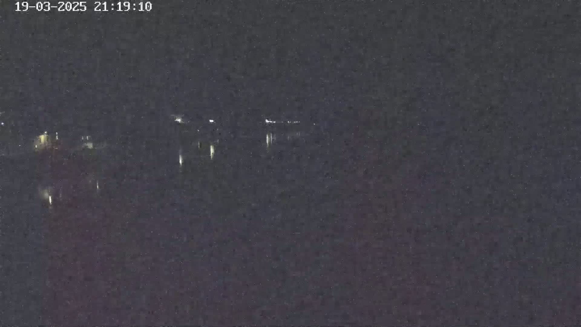 A nighttime long shot shows a dark body of water reflecting a few distant lights under a clear, dark sky.