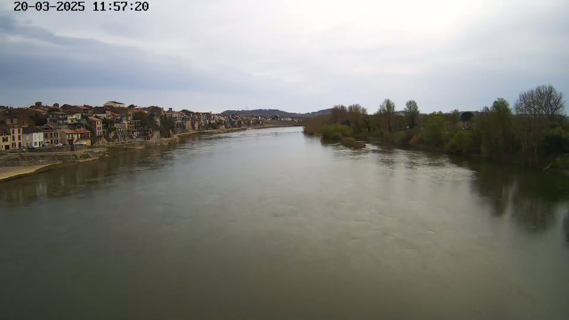 A wide, calm river flows past a town of closely-packed buildings under a cloudy sky.