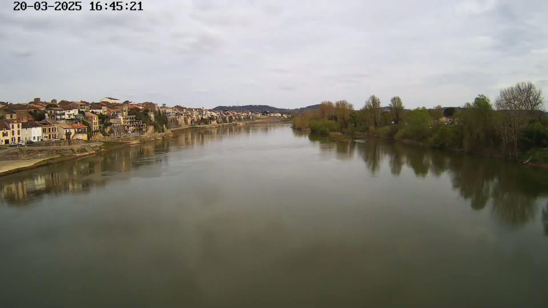 A wide, calm river reflects the overcast sky and the buildings and trees lining its banks.