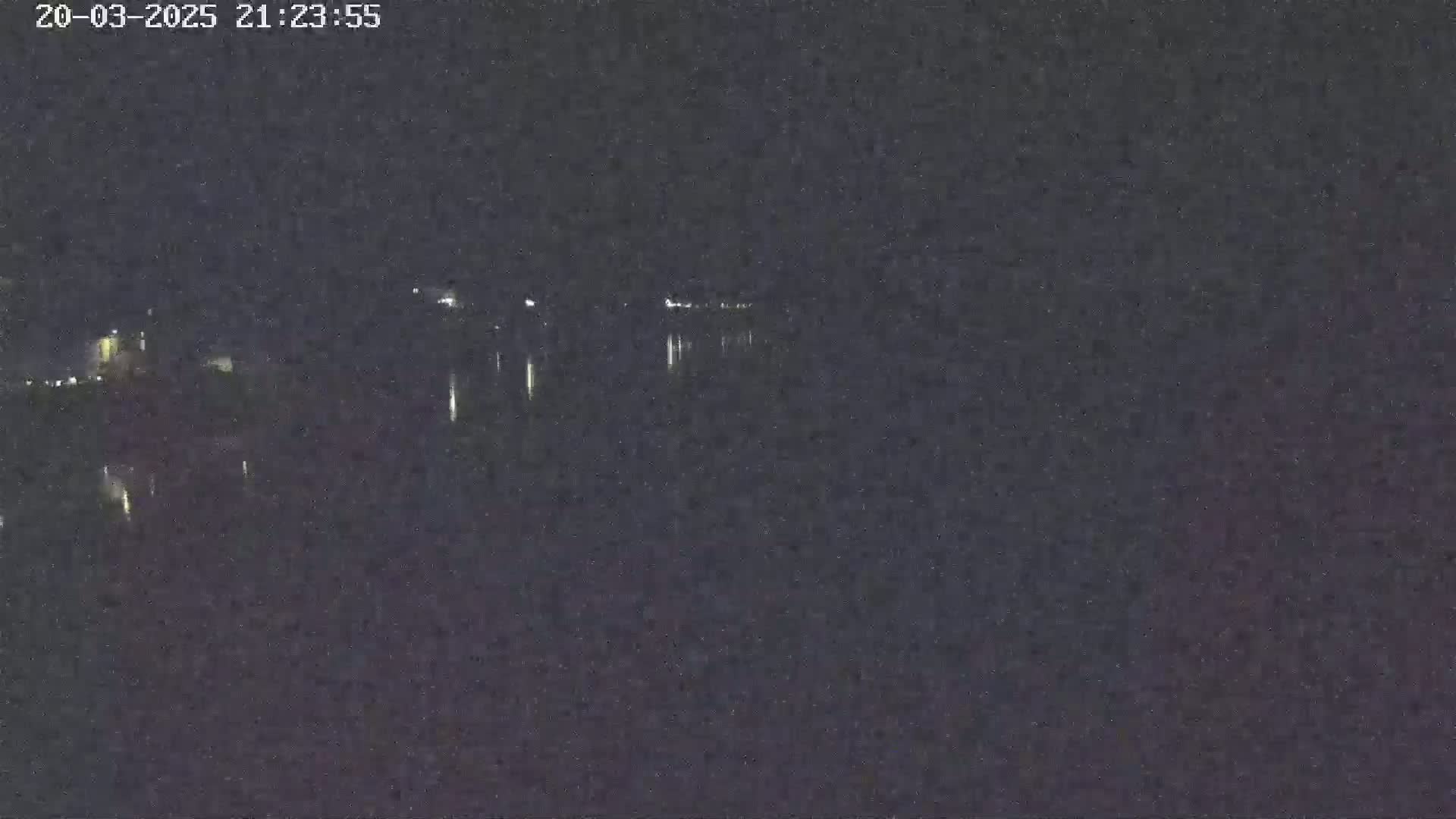 A dark nighttime scene shows a body of water reflecting faint lights from a distant shoreline under a clear sky.