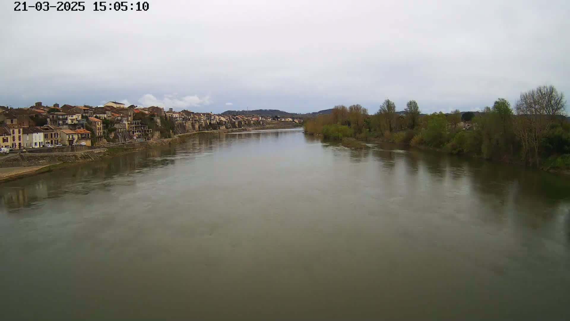 A wide, calm river flows past a village of closely-packed buildings under an overcast sky.