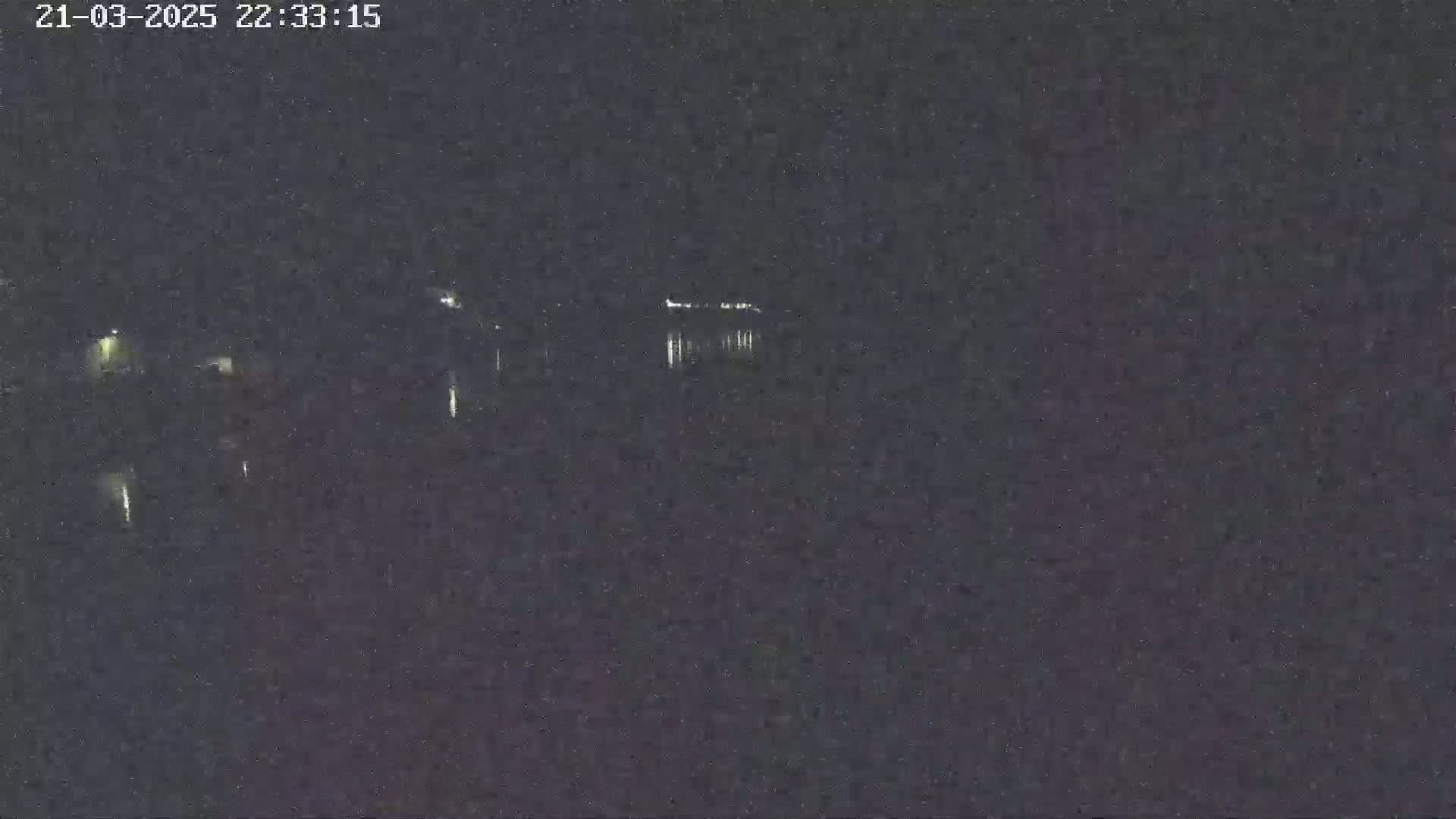 A nighttime, low-light image shows a dark body of water reflecting a few distant lights under a dark sky.