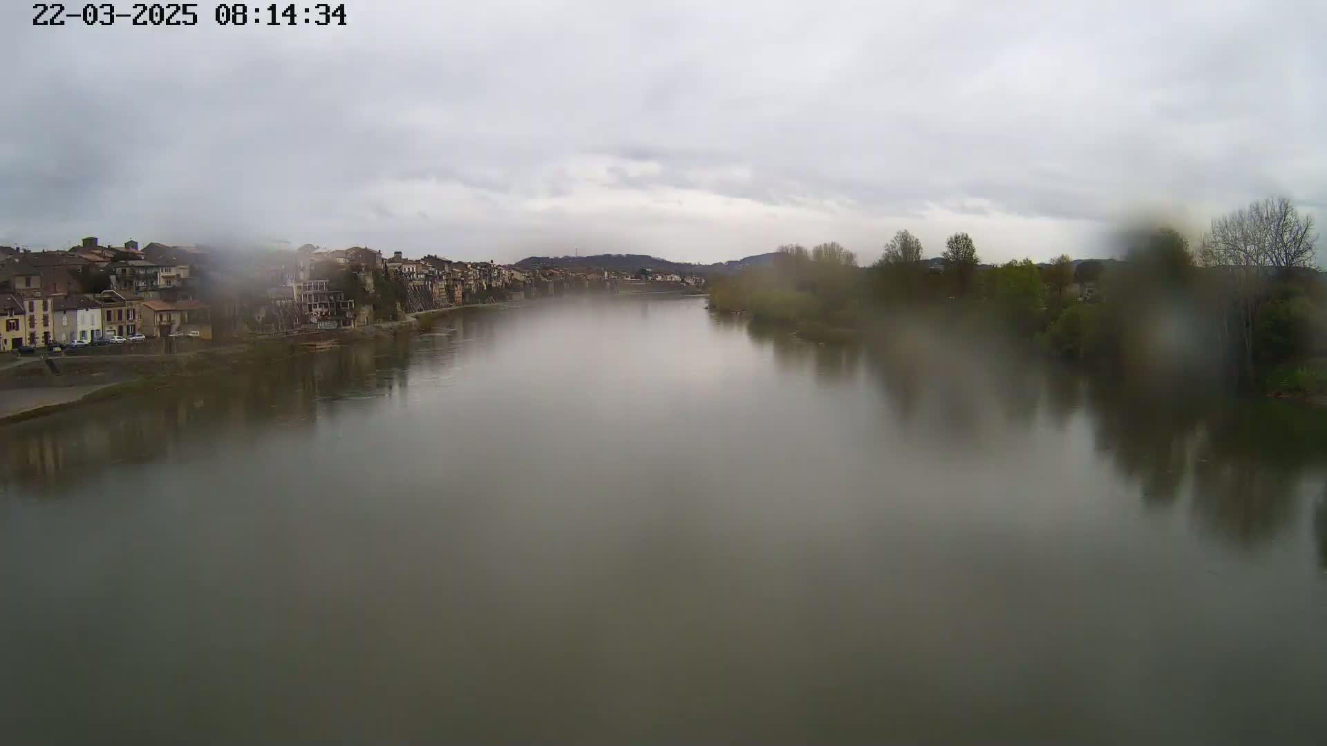 A wide, calm river is seen on a cloudy, rainy day, with a town on one bank and trees on the other.