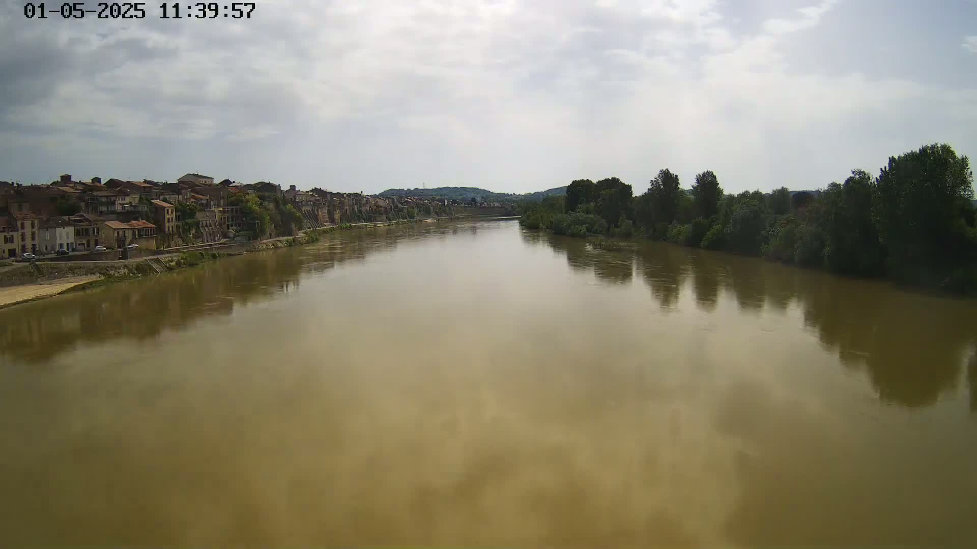 A wide, muddy river flows past a town under a mostly cloudy sky.
