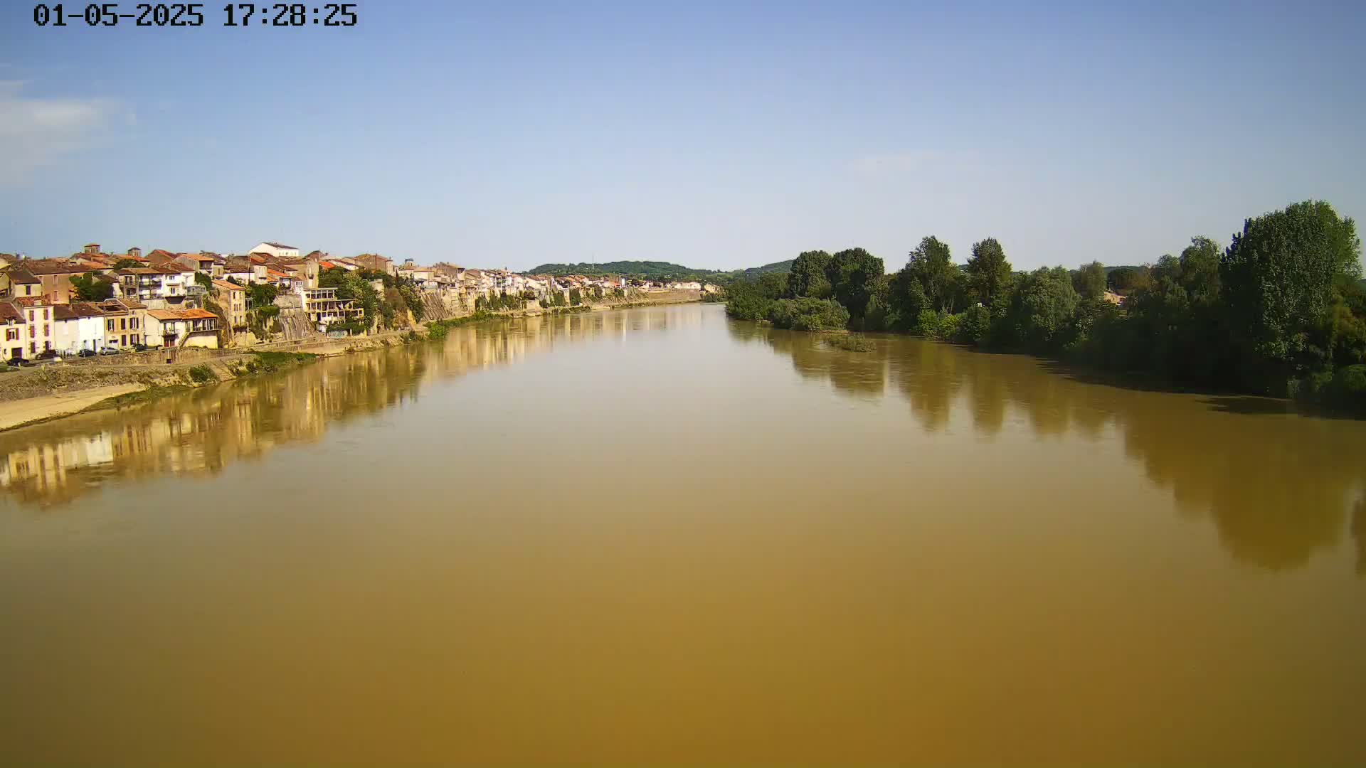 A wide, muddy river reflects a line of buildings and trees under a clear, bright sky.
