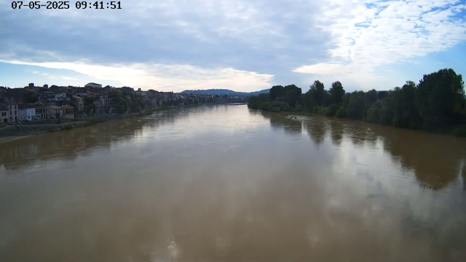 A wide, muddy river reflects a partly cloudy sky, with a town on one bank and trees lining the opposite bank.