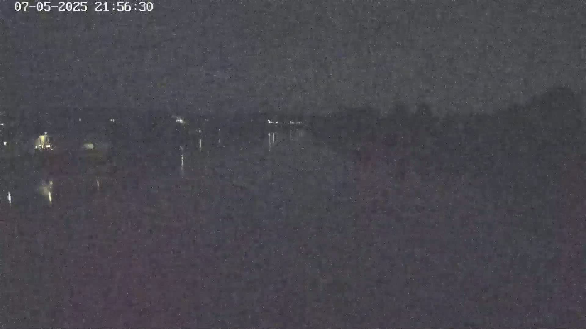 A nighttime, low-light view shows a dark body of water reflecting faint lights from the opposite shore.